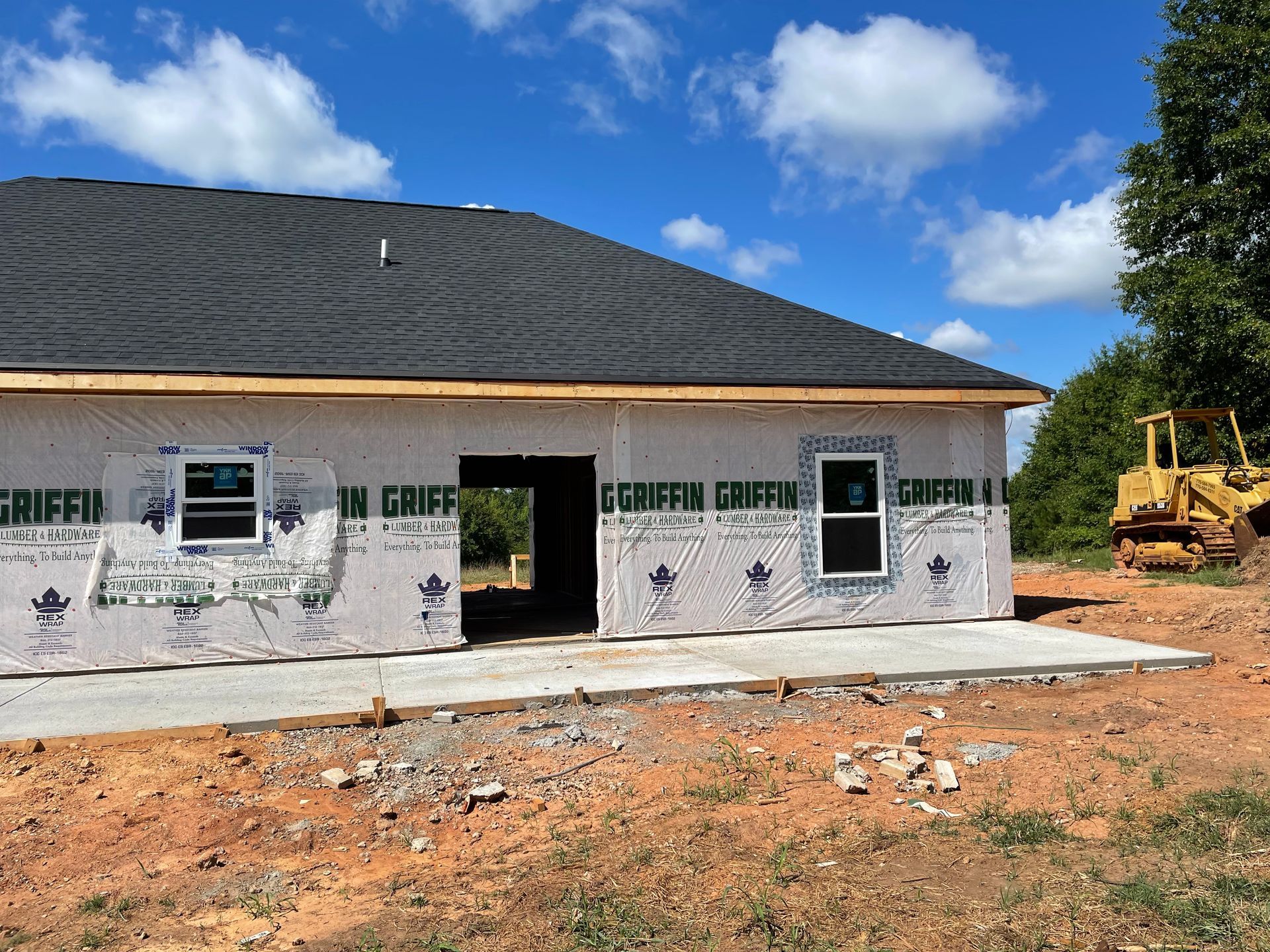 Partially built house with white wrap, dark roof, and construction equipment on a dirt lot under a blue sky