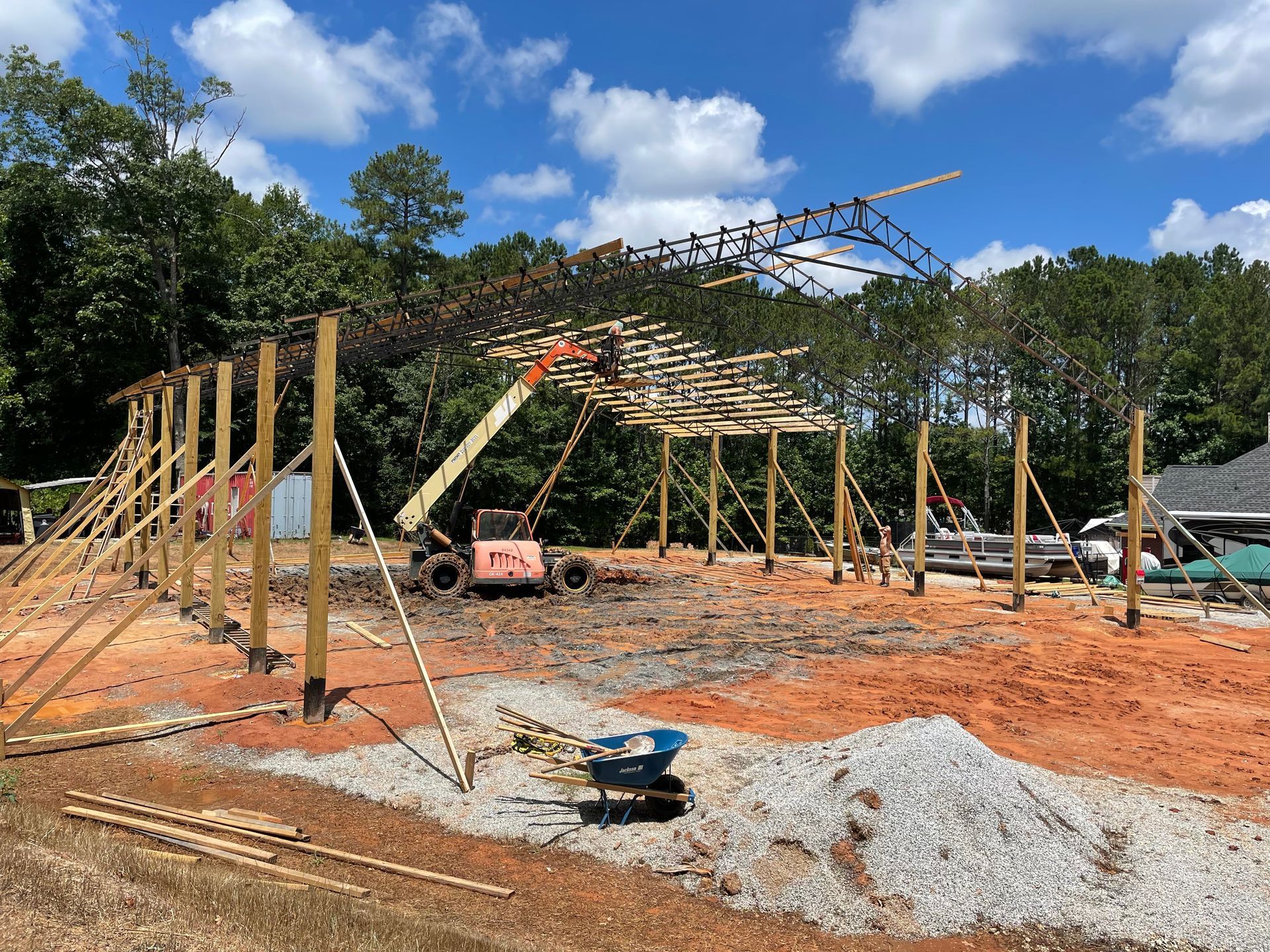 Steel frame of a building under construction on a dirt lot, with workers and trees under a blue sky.
