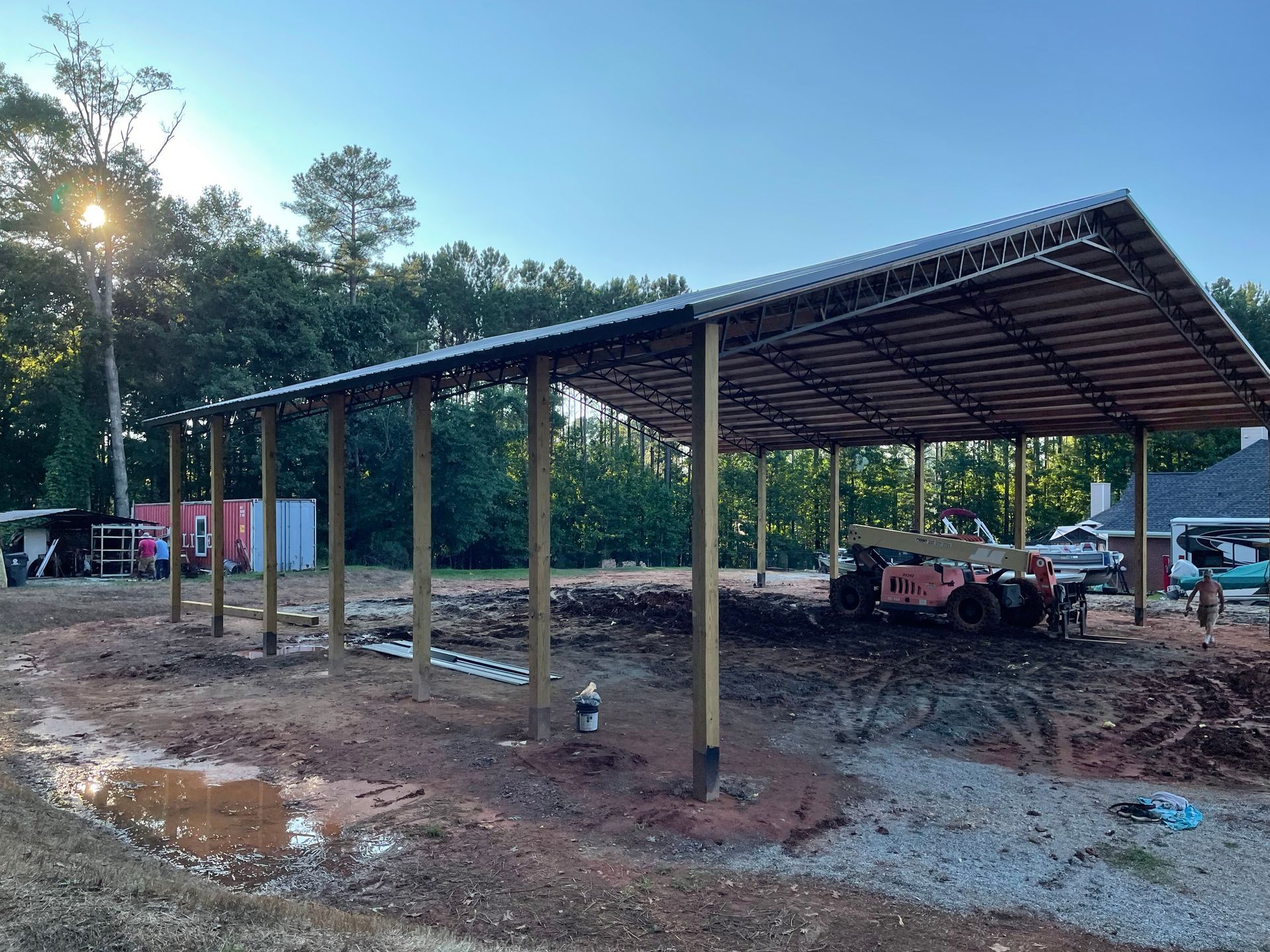 Open-sided metal shed in a muddy clearing, with trees and vehicles in the background