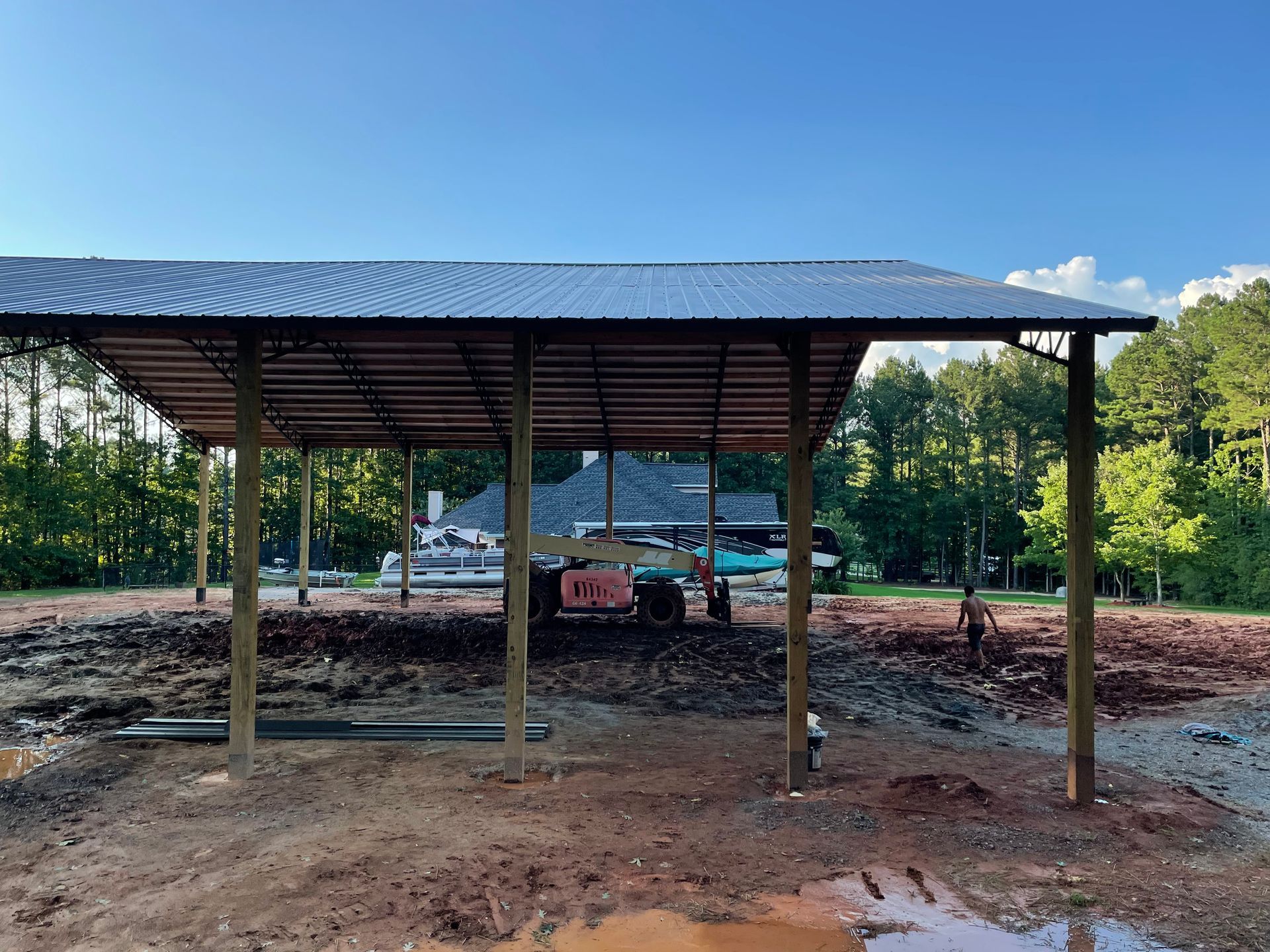 Open-sided metal shelter on a dirt lot with a parked car and trees in the background