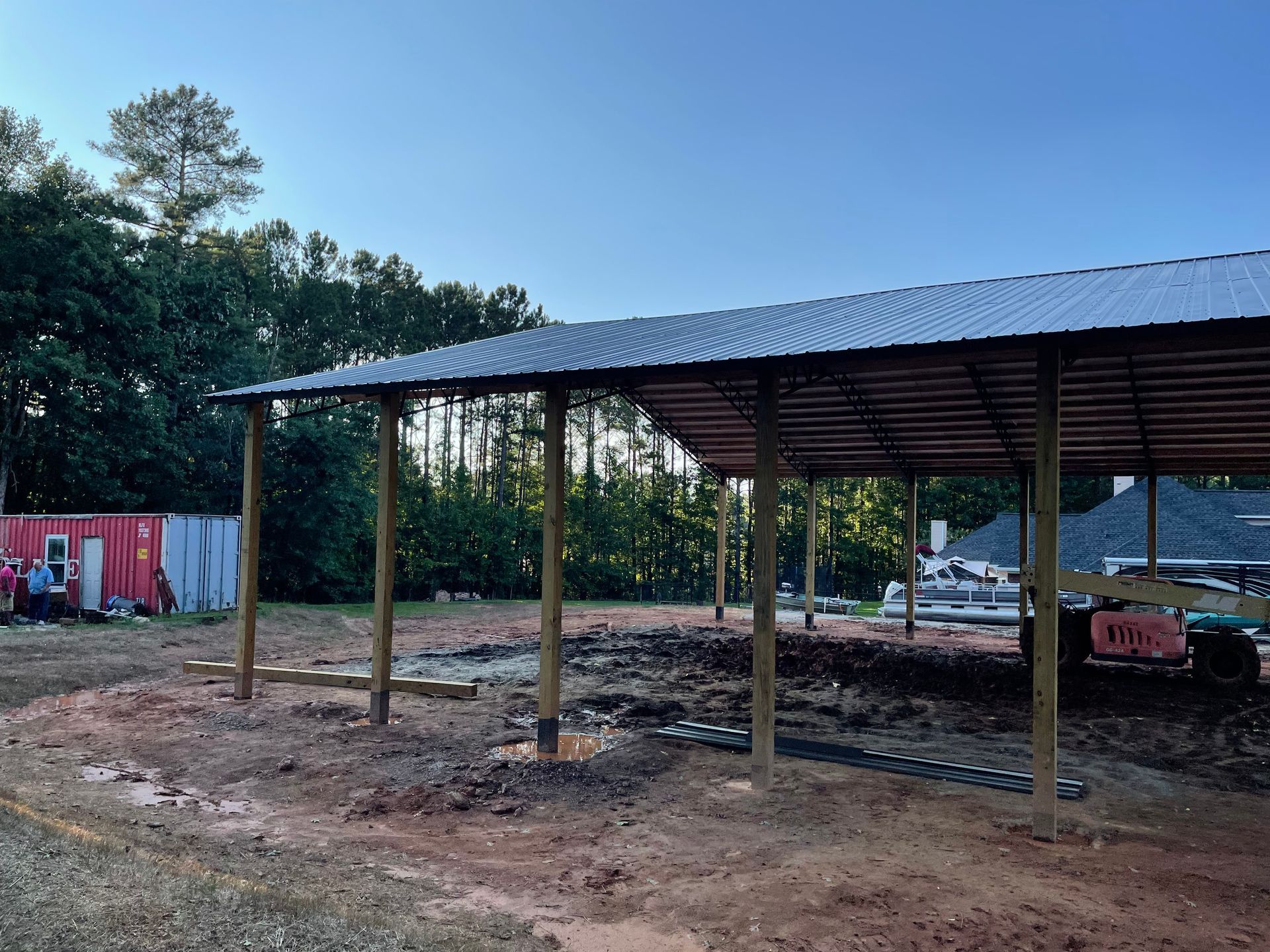 Open-sided metal-roof shelter on posts in a dirt yard, with trees and a parked tractor nearby.