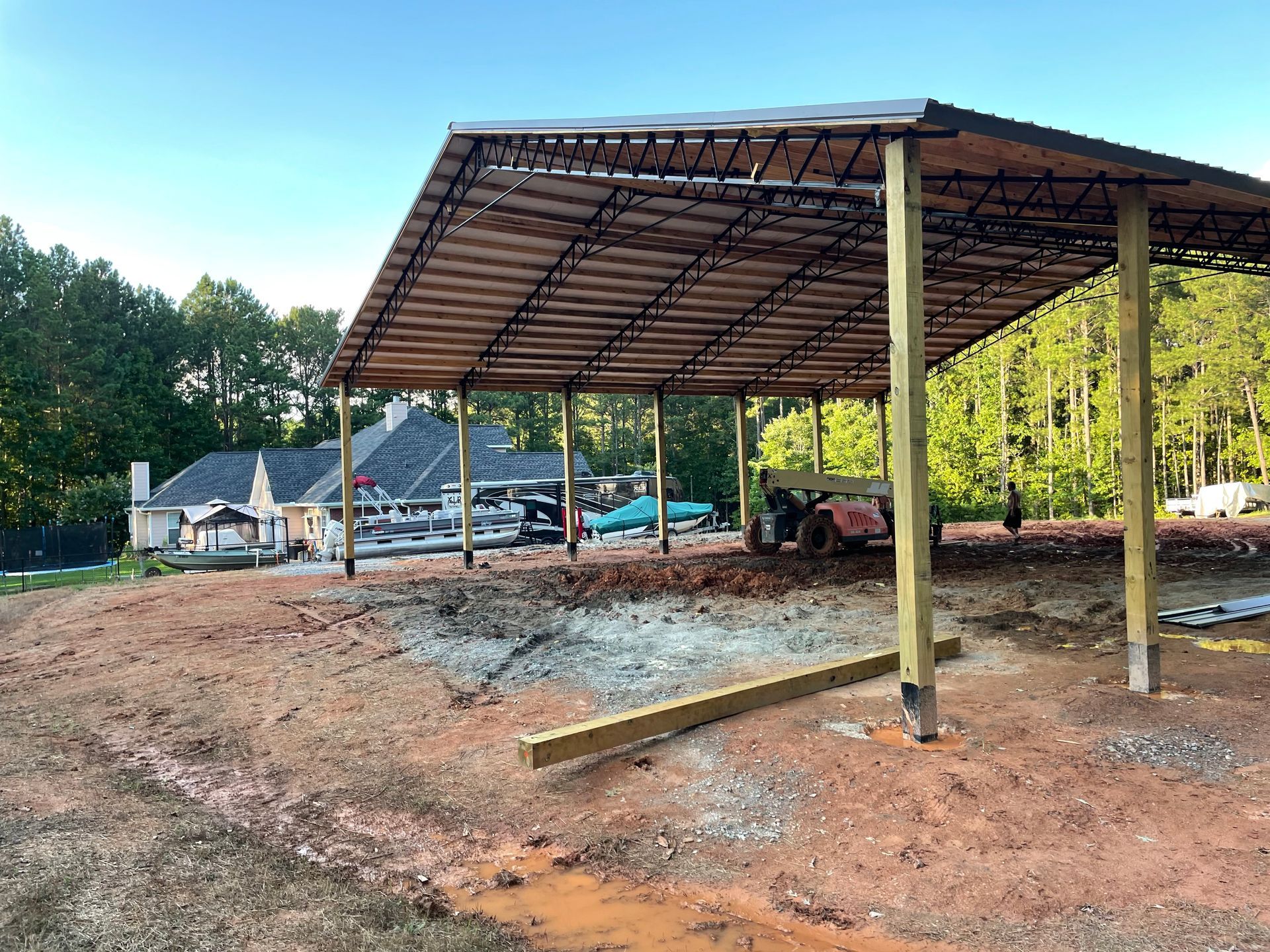Partially built metal pavilion roof over a dirt lot with construction equipment and trees nearby