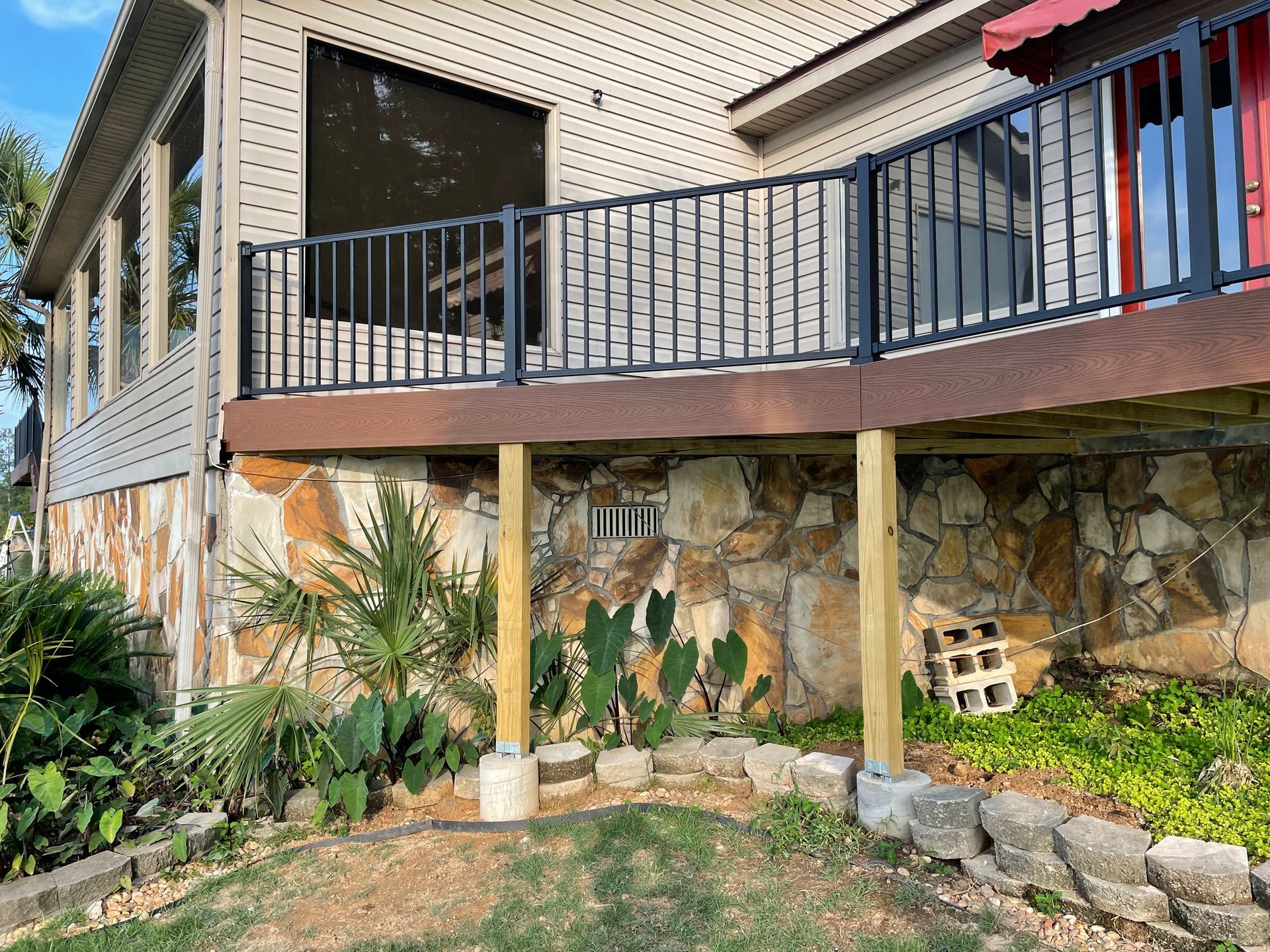 House exterior with raised deck, black railing, stone foundation, and landscaped plants underneath