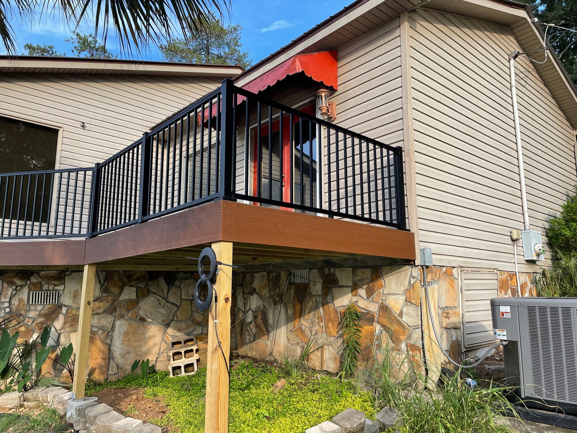 House deck with black railing, red awning, and stone foundation beside an HVAC unit