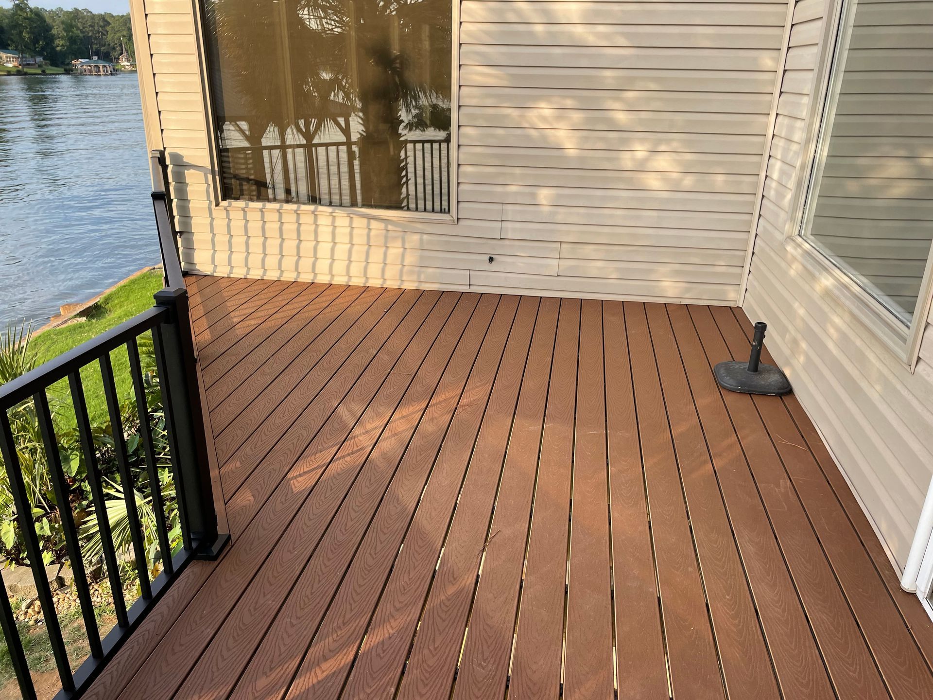 Wooden deck beside a house, overlooking water, with black railing and sunlight on the siding.