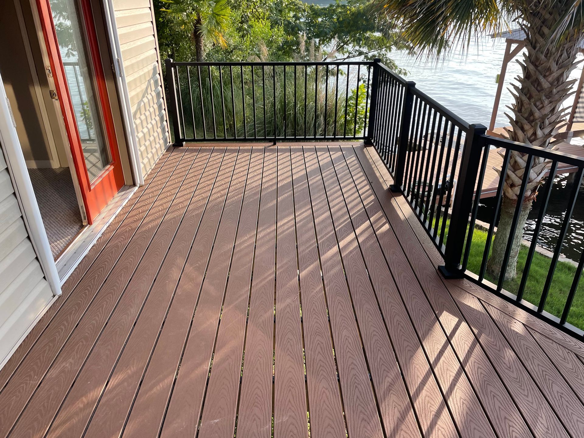 Wooden balcony deck with black railings, next to a glass door and overlooking trees and water.