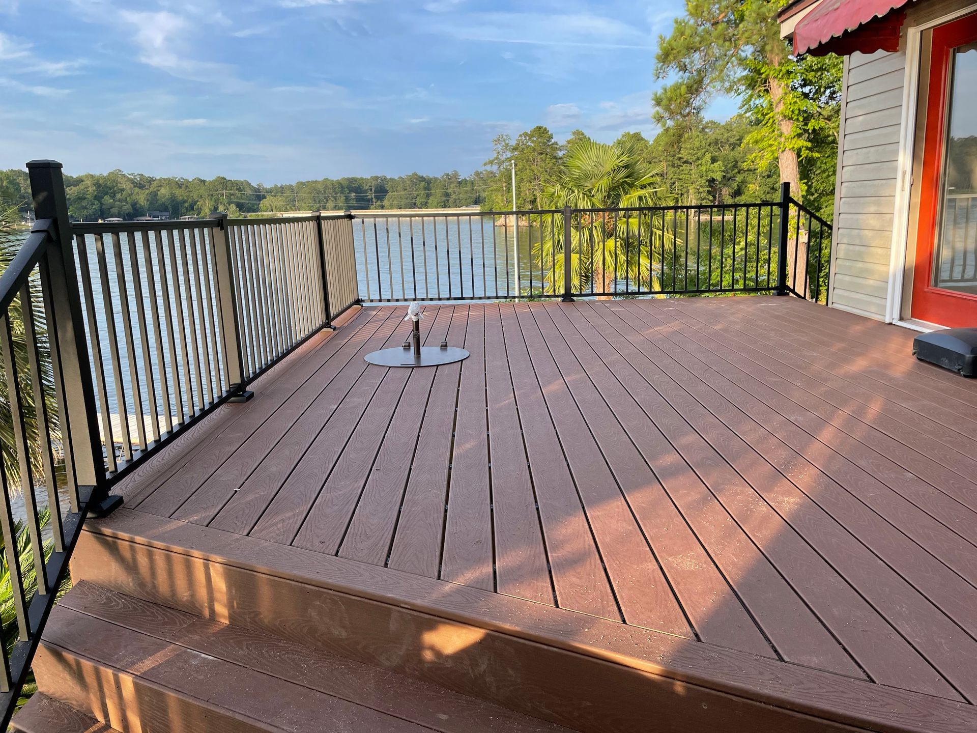 Wooden deck with black railing overlooking a lake and trees, beside a red house