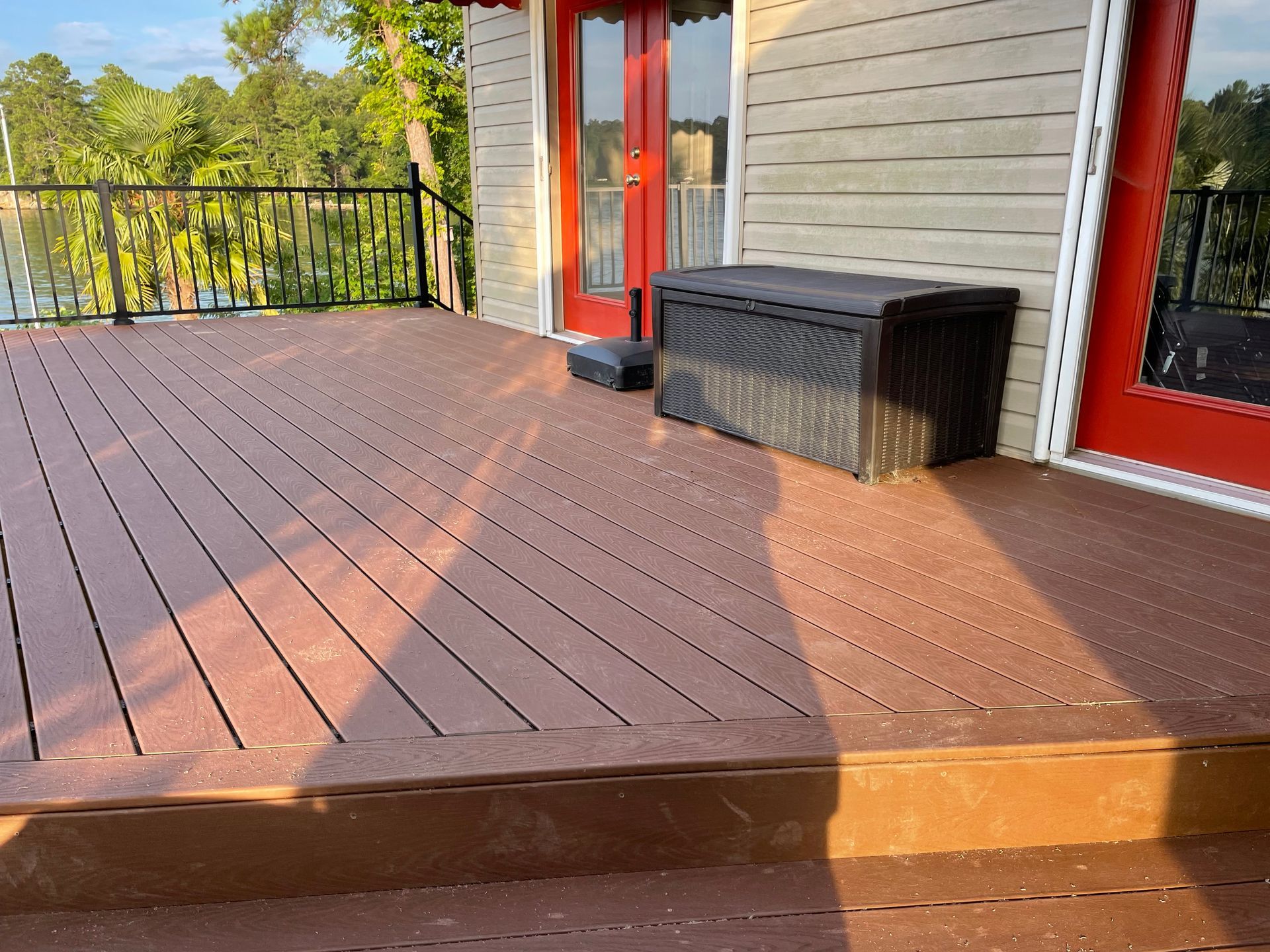 Wooden deck with black railing, red doors, and a wicker storage bench beside a house.
