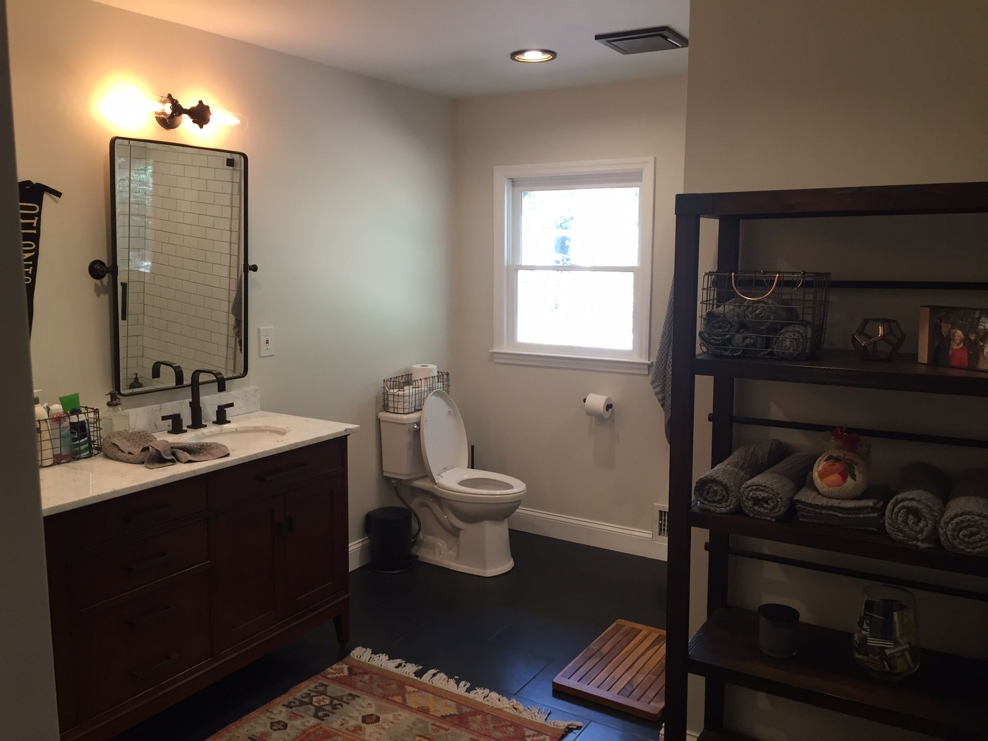 Bathroom with dark vanity, toilet, window, and shelving unit on a blue rug