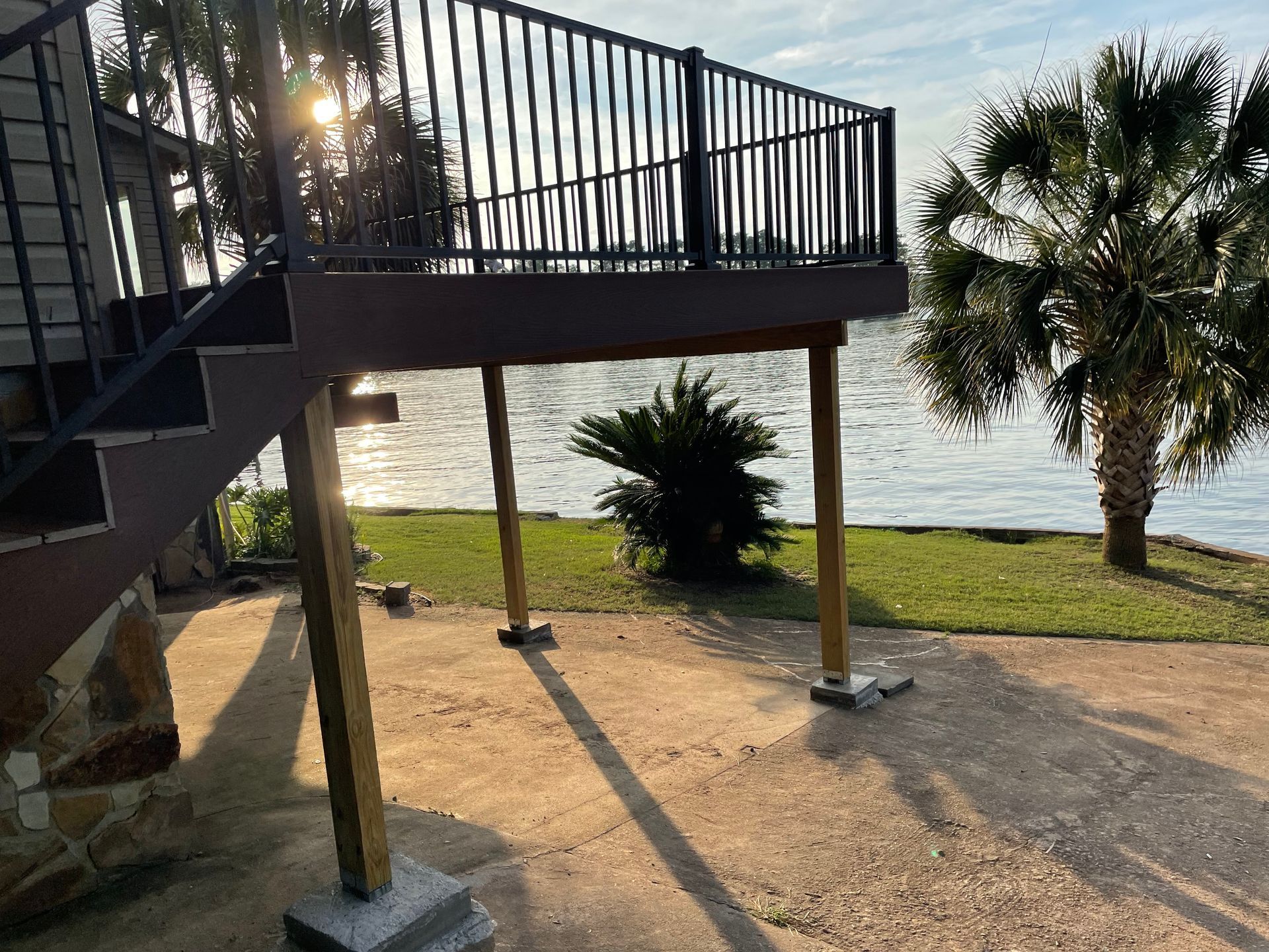 Covered patio deck by a lake with stairs, railings, and palm trees at sunset.