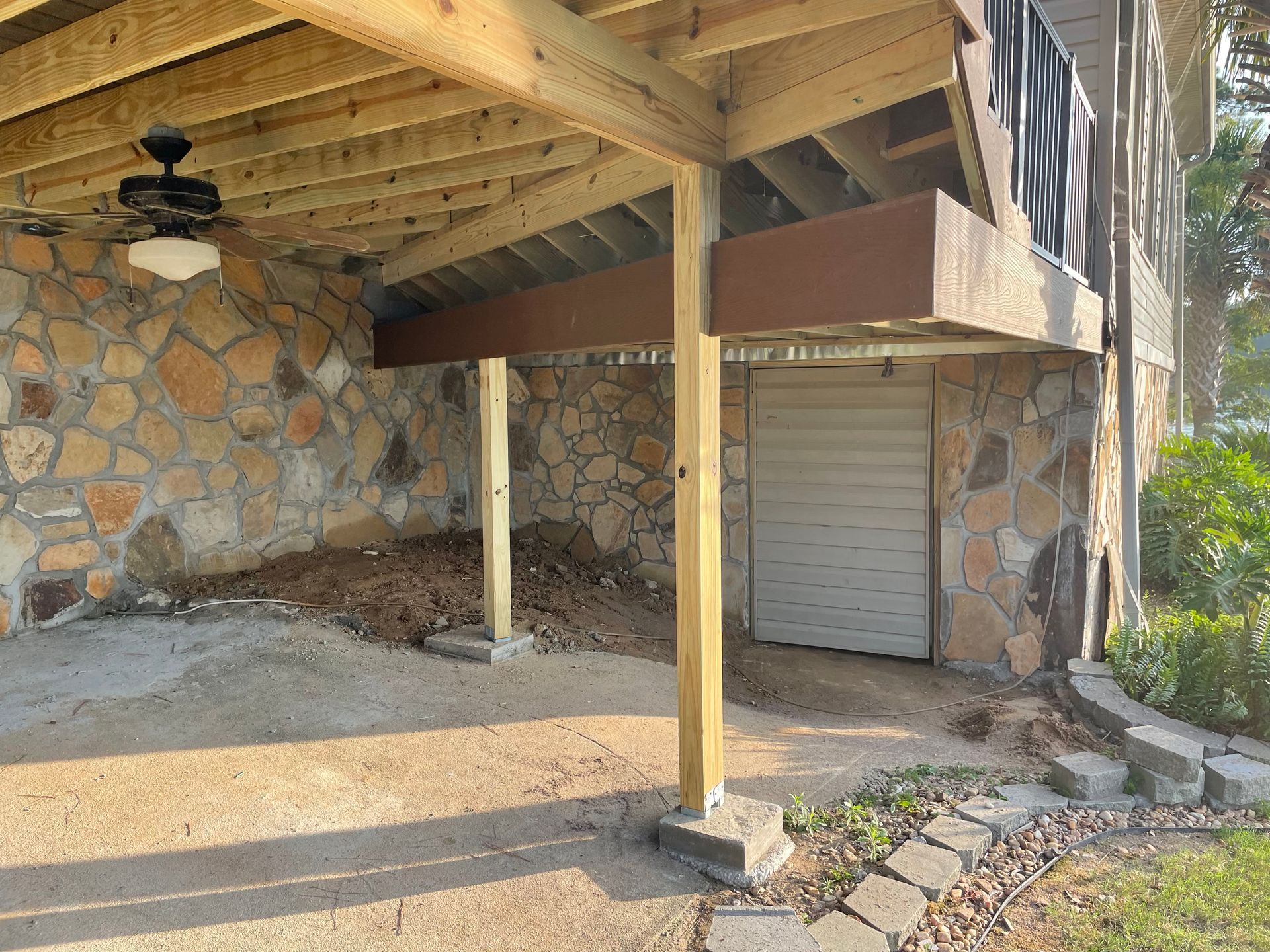 Covered stone patio with wooden beams, ceiling fan, and a closed gray door under a raised deck