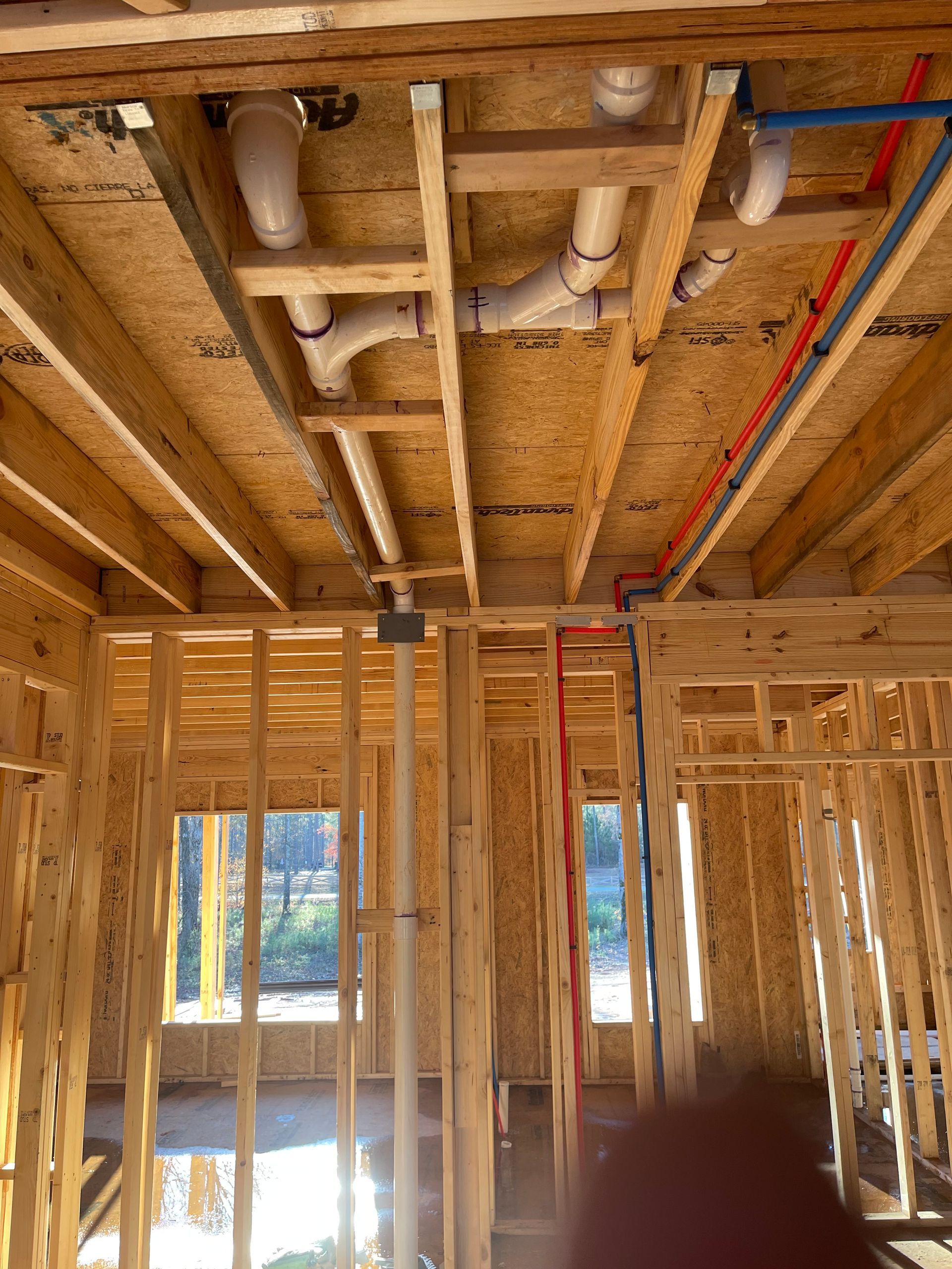 Interior of a wood-framed house under construction with exposed plumbing and wiring in the ceiling.