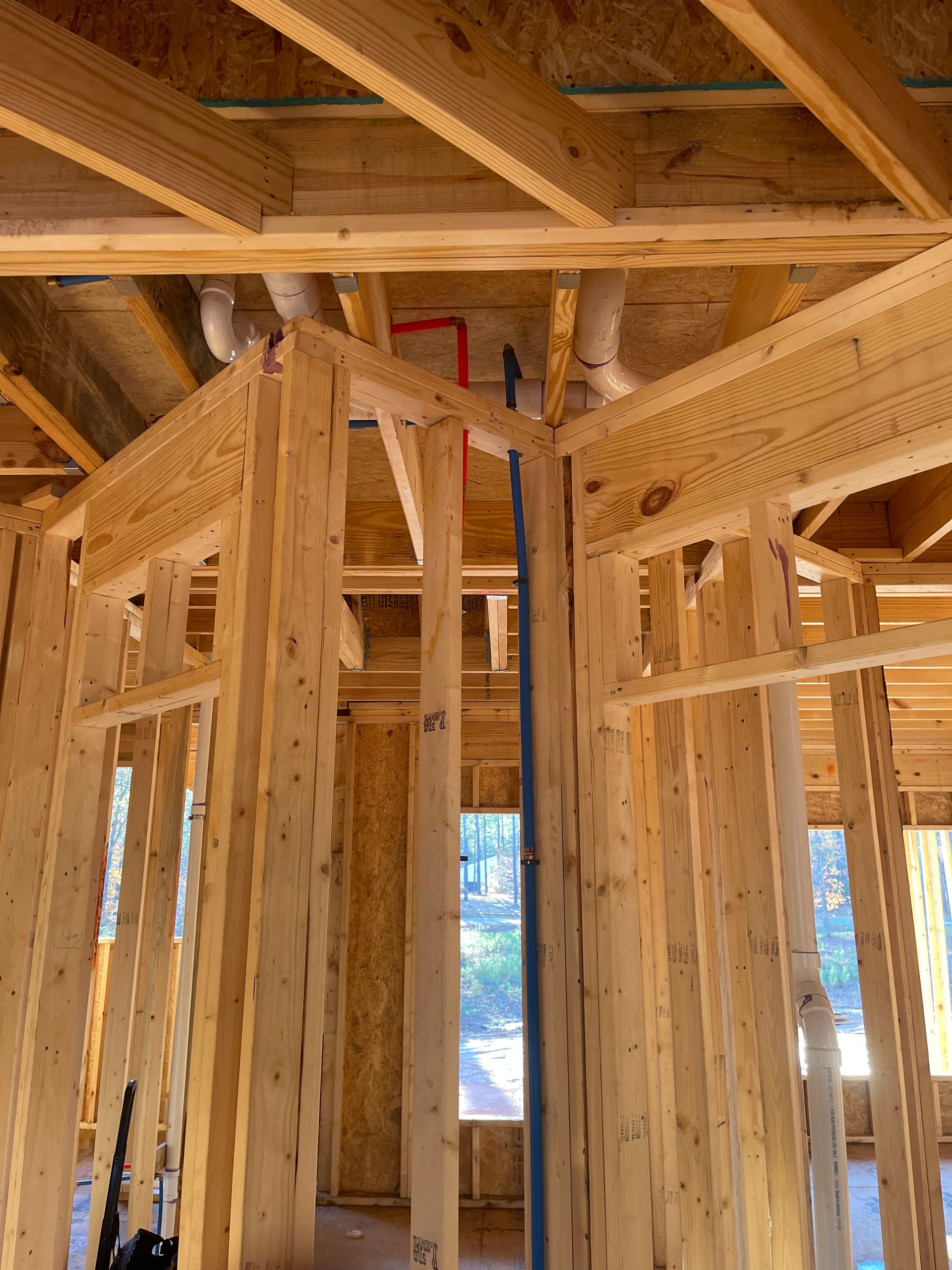 Interior of a wood-framed house under construction, showing exposed studs, beams, and window openings.