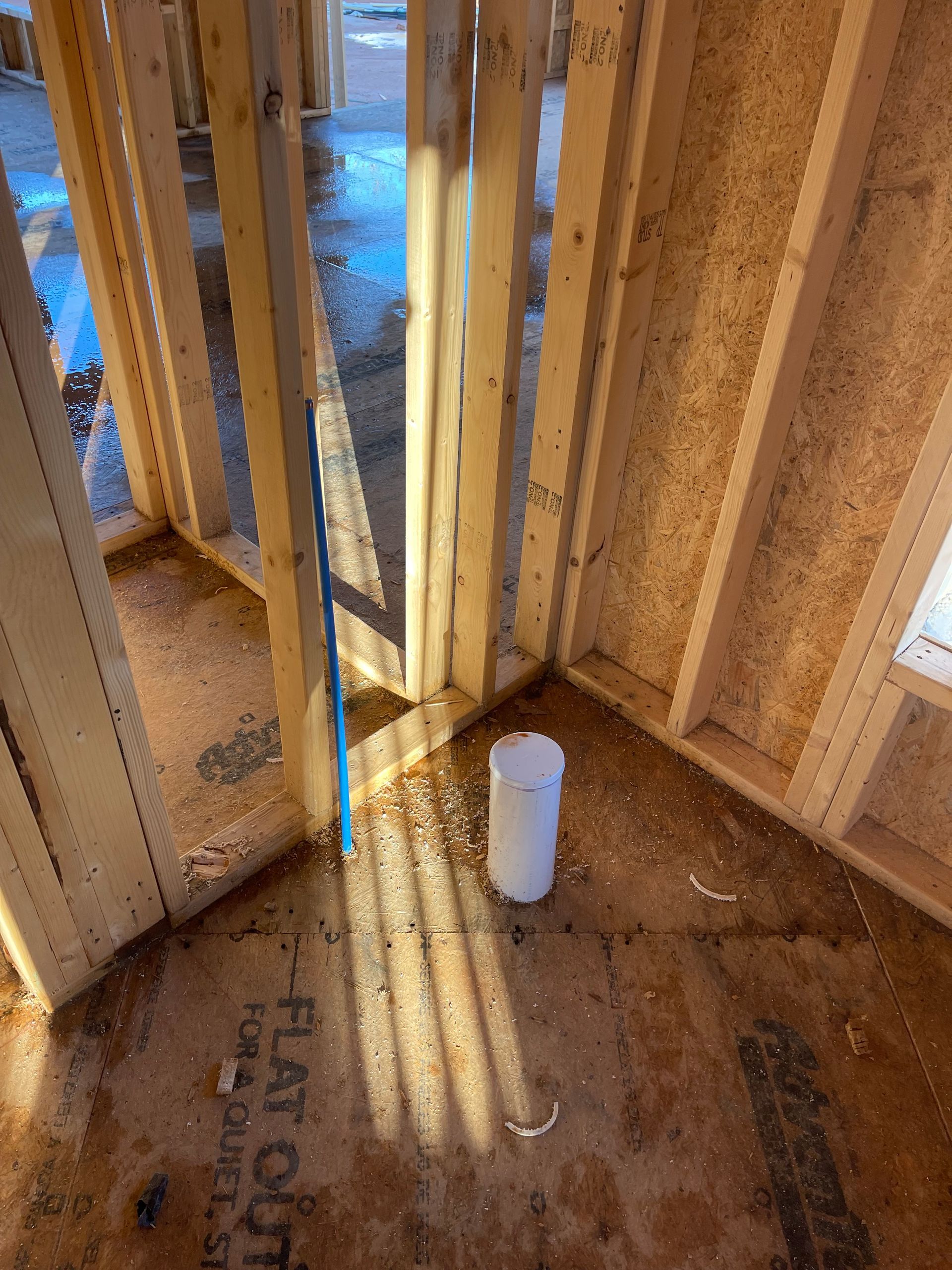 Partially framed wall corner with sunlight casting shadows on plywood floor and a white pipe stump nearby