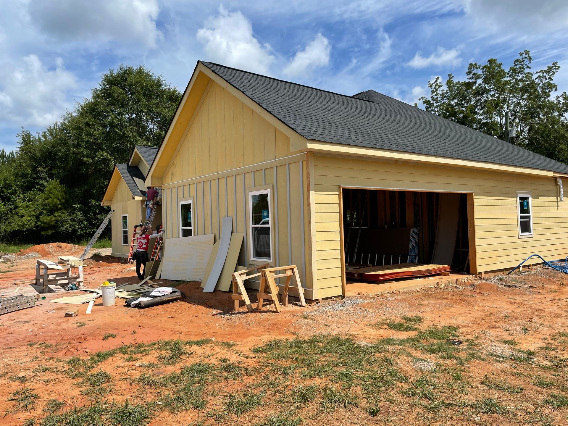 Yellow house under construction with open garage, workers, and building materials on a dirt lot