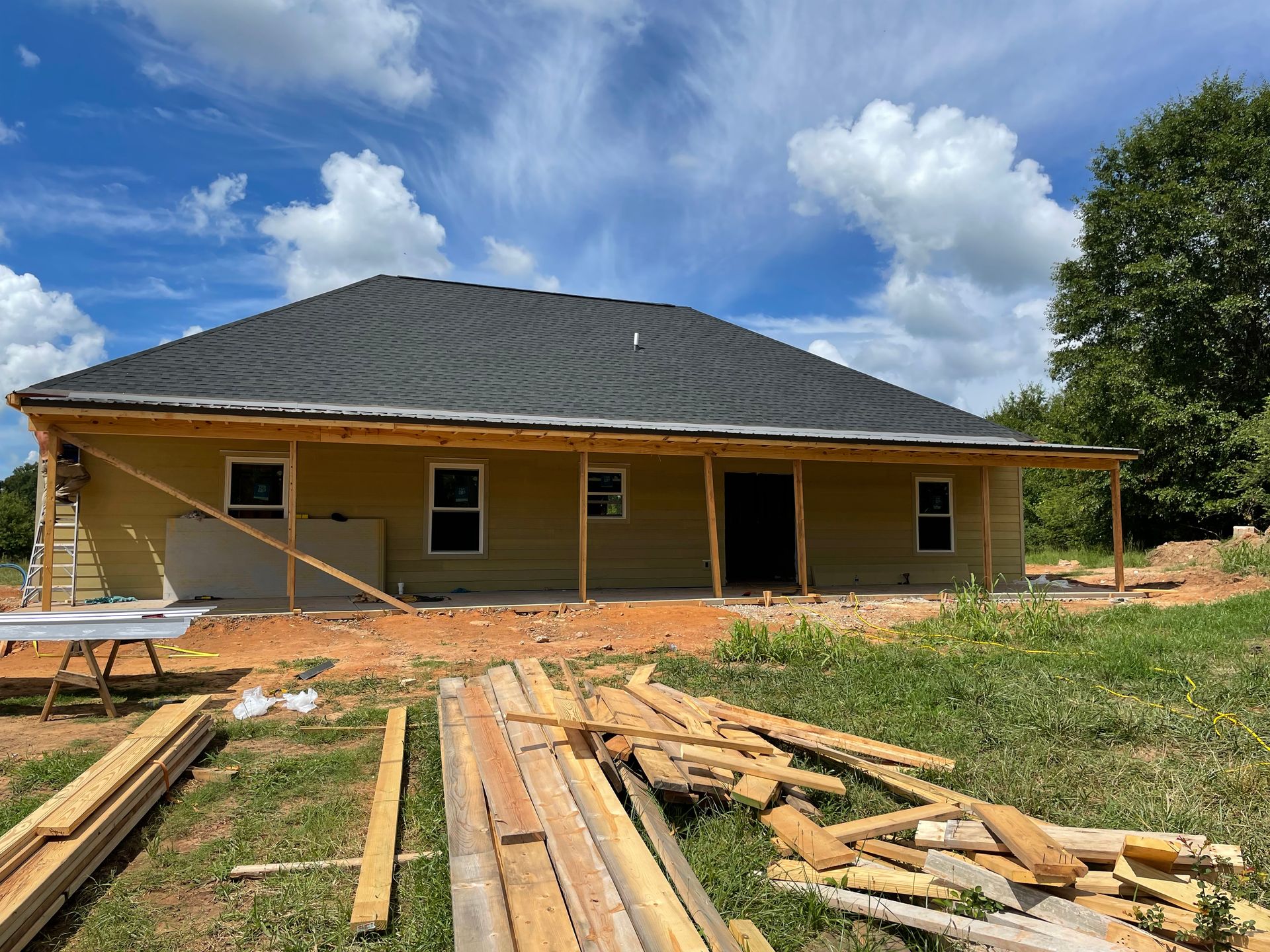 Yellow house under construction with black roof, lumber in the foreground, and a blue sky with clouds
