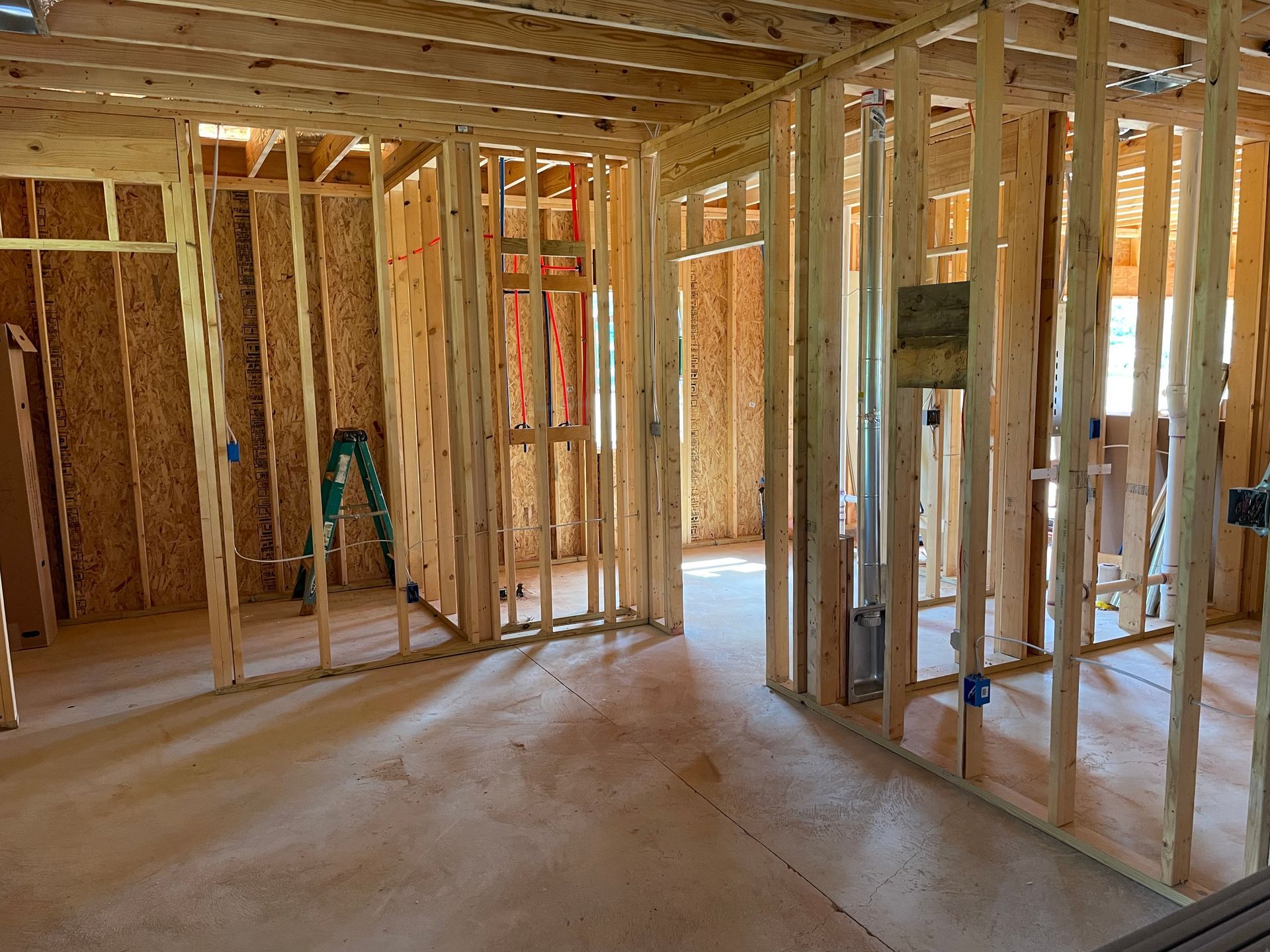Unfinished house interior with exposed wooden framing, drywall, and a ladder on a concrete floor.