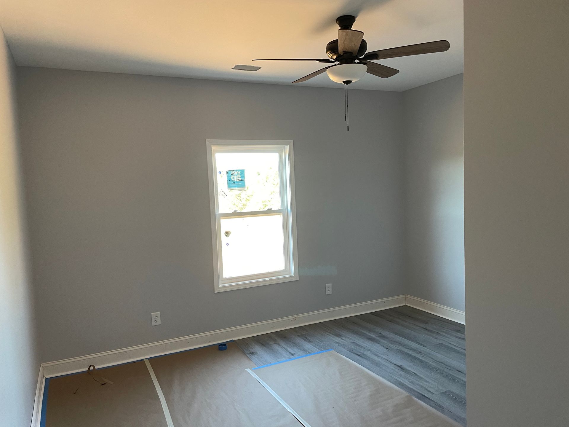 Empty gray bedroom with a window, ceiling fan, and new flooring partially installed