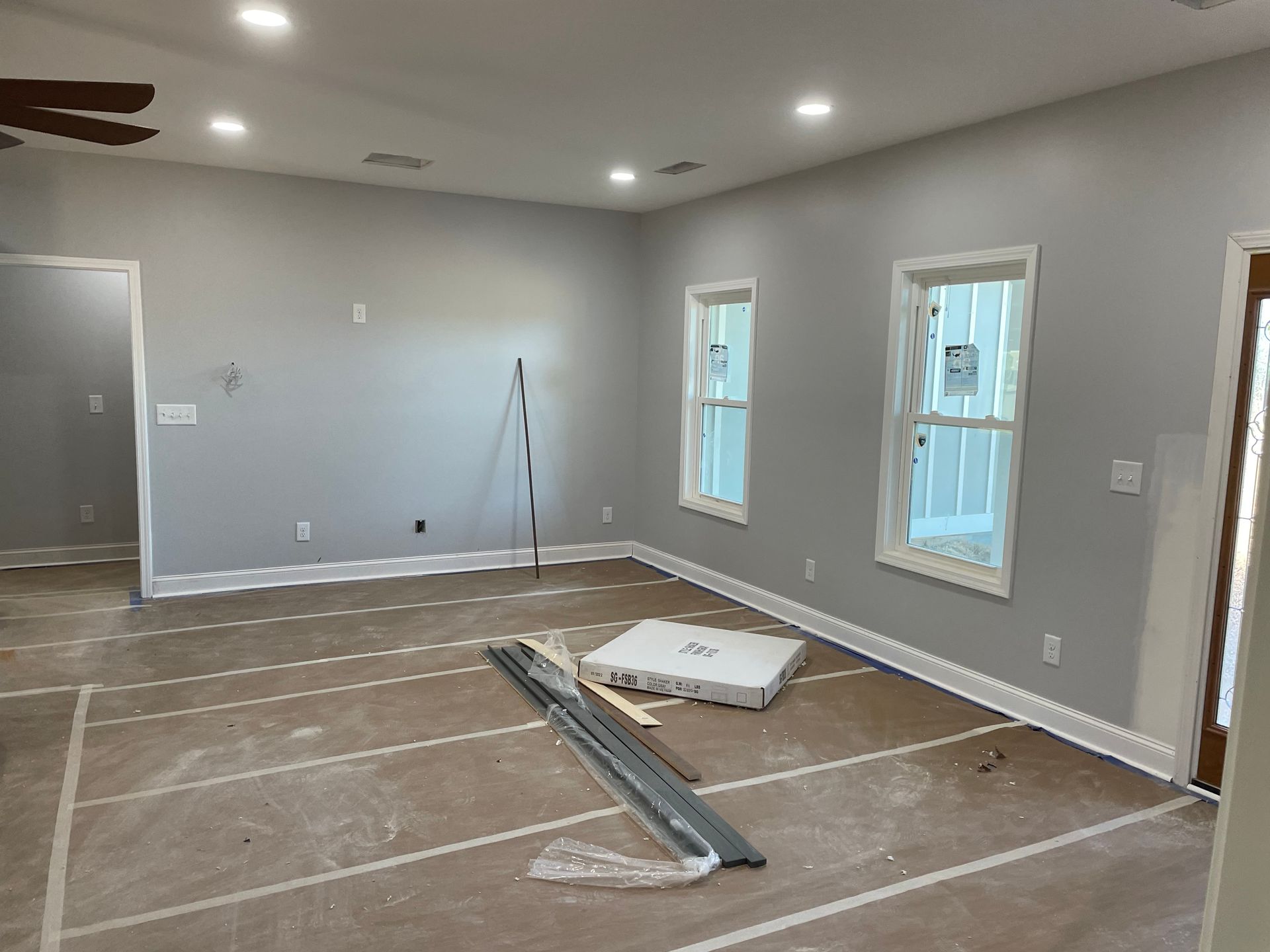 Empty gray room under renovation with flooring materials and tools on the floor, two windows on the right.