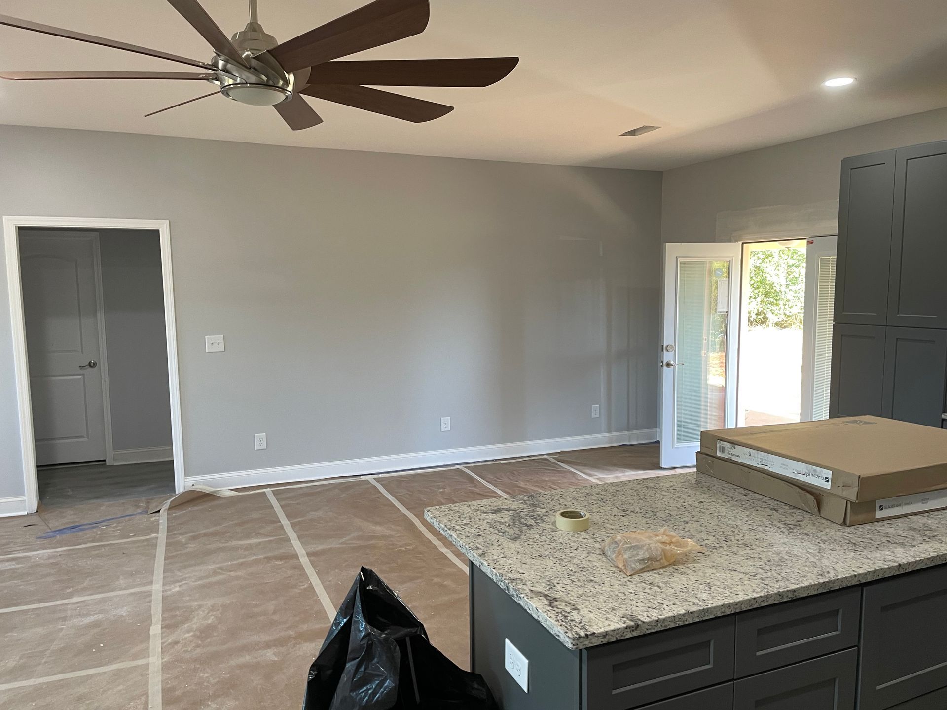 Empty living room under renovation with gray walls, exposed flooring, ceiling fan, and kitchen island in foreground
