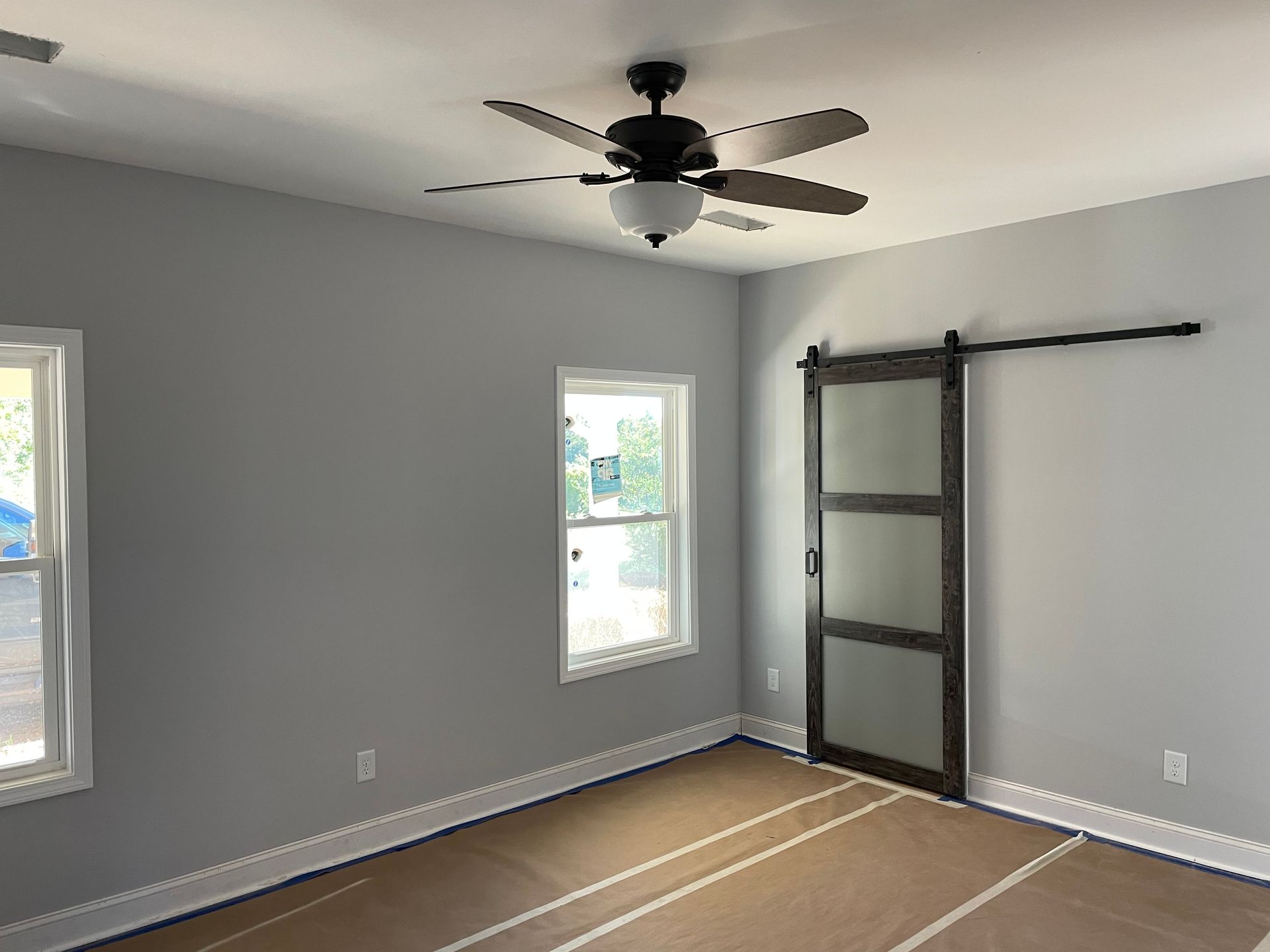 Empty gray bedroom with three windows, ceiling fan, and black wall-mounted TV bracket on the right.