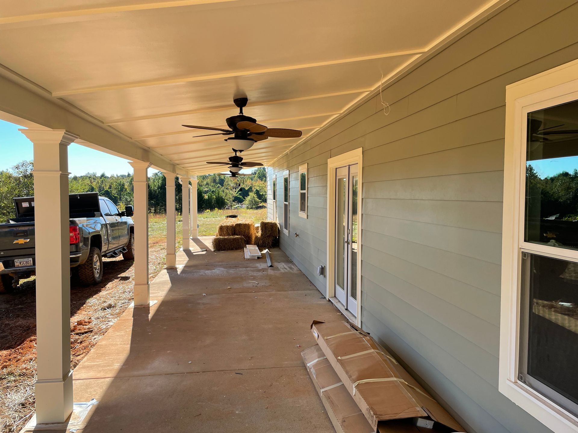 Covered front porch with light gray siding, ceiling fans, windows, and construction materials on the floor.