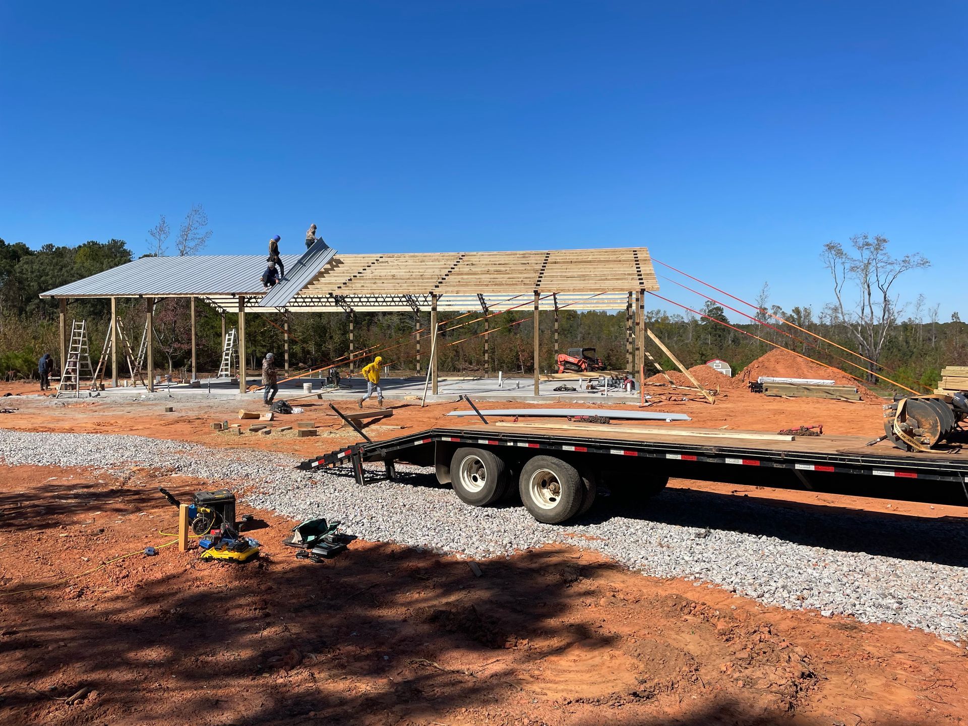 Construction site with a partially built metal roof frame, gravel foreground, and trailer under a clear blue sky