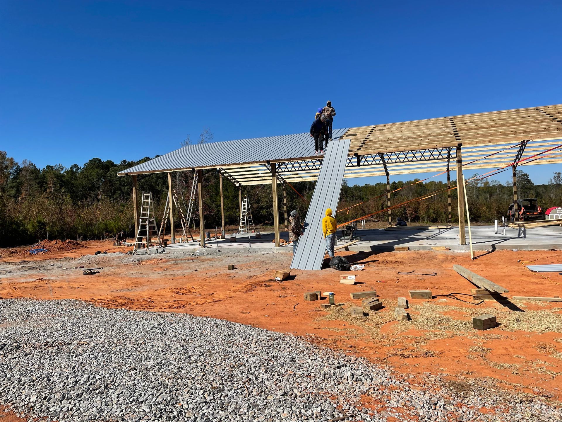 Construction site with a metal frame building, worker on a ladder, and gravel and dirt foreground under a blue sky
