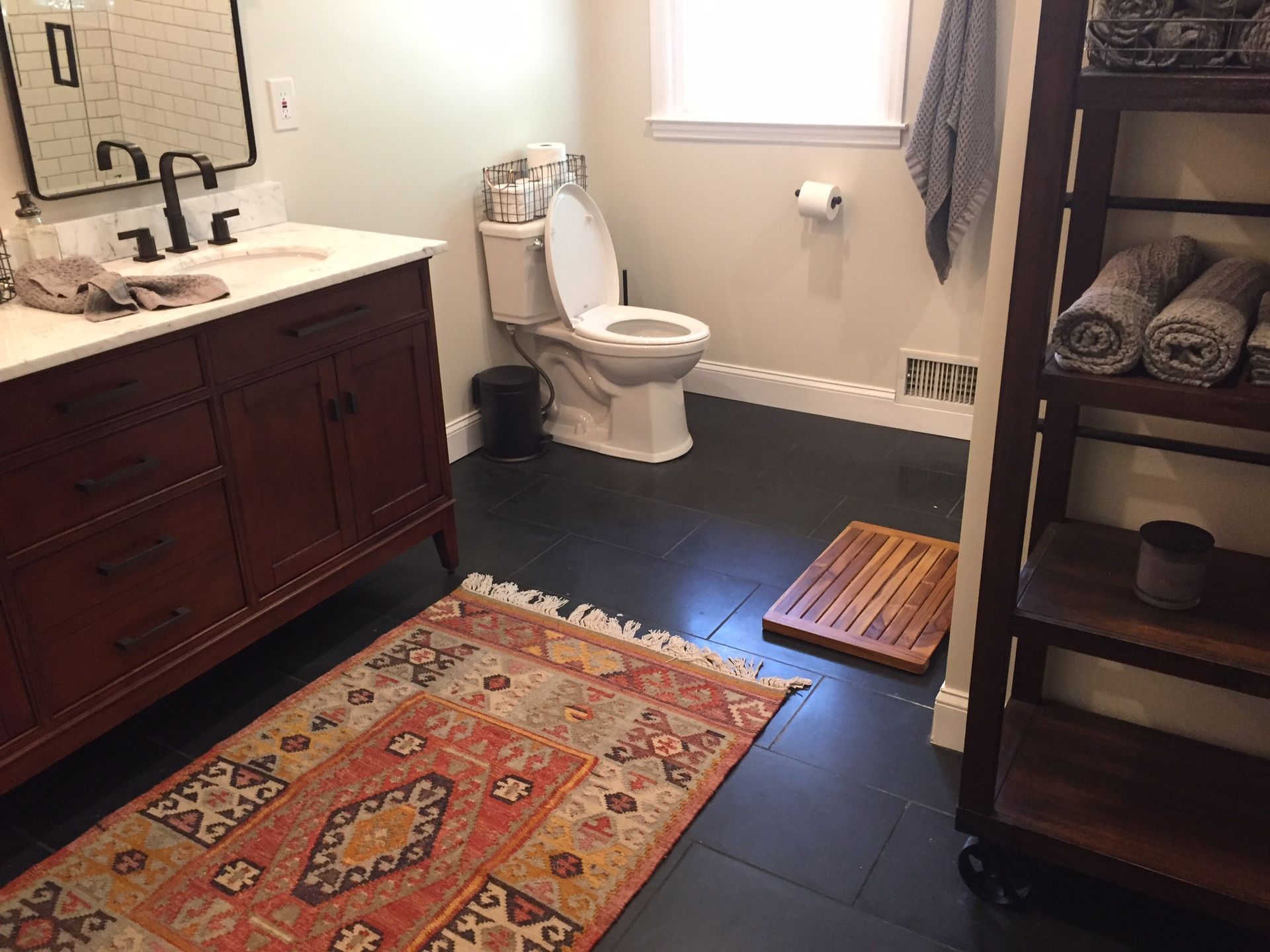 Bathroom with dark vanity, white toilet, patterned rug, and shelving unit with towels.