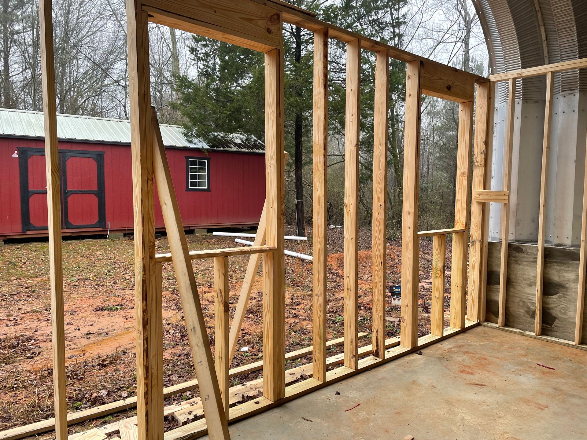 Exposed wooden wall framing inside a shed or barn, with a red building visible outside.