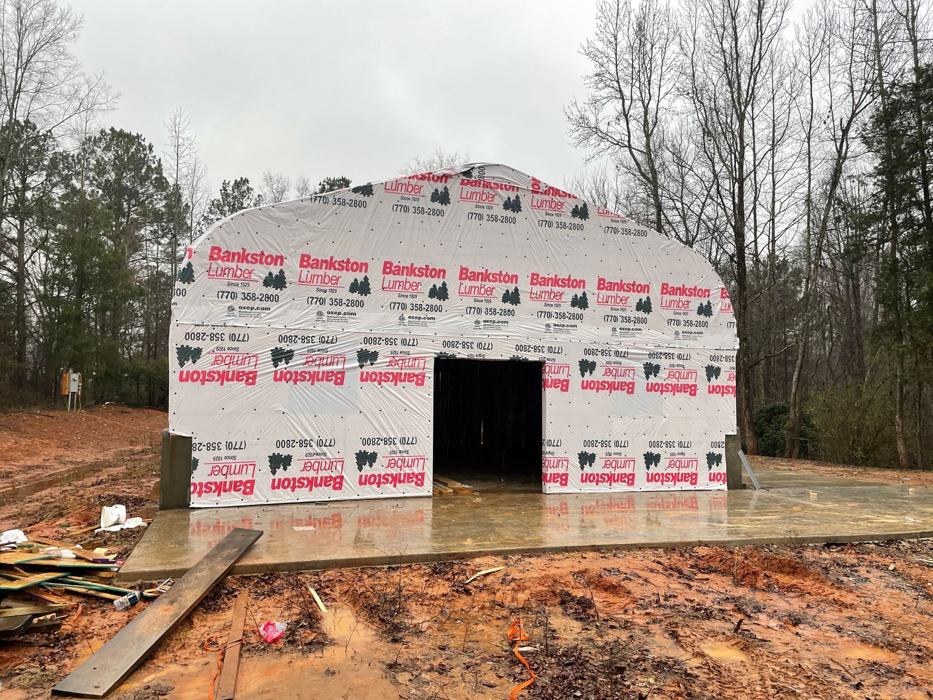 Partially built house framed in white weatherproof wrap, with an open doorway on a muddy wooded lot.