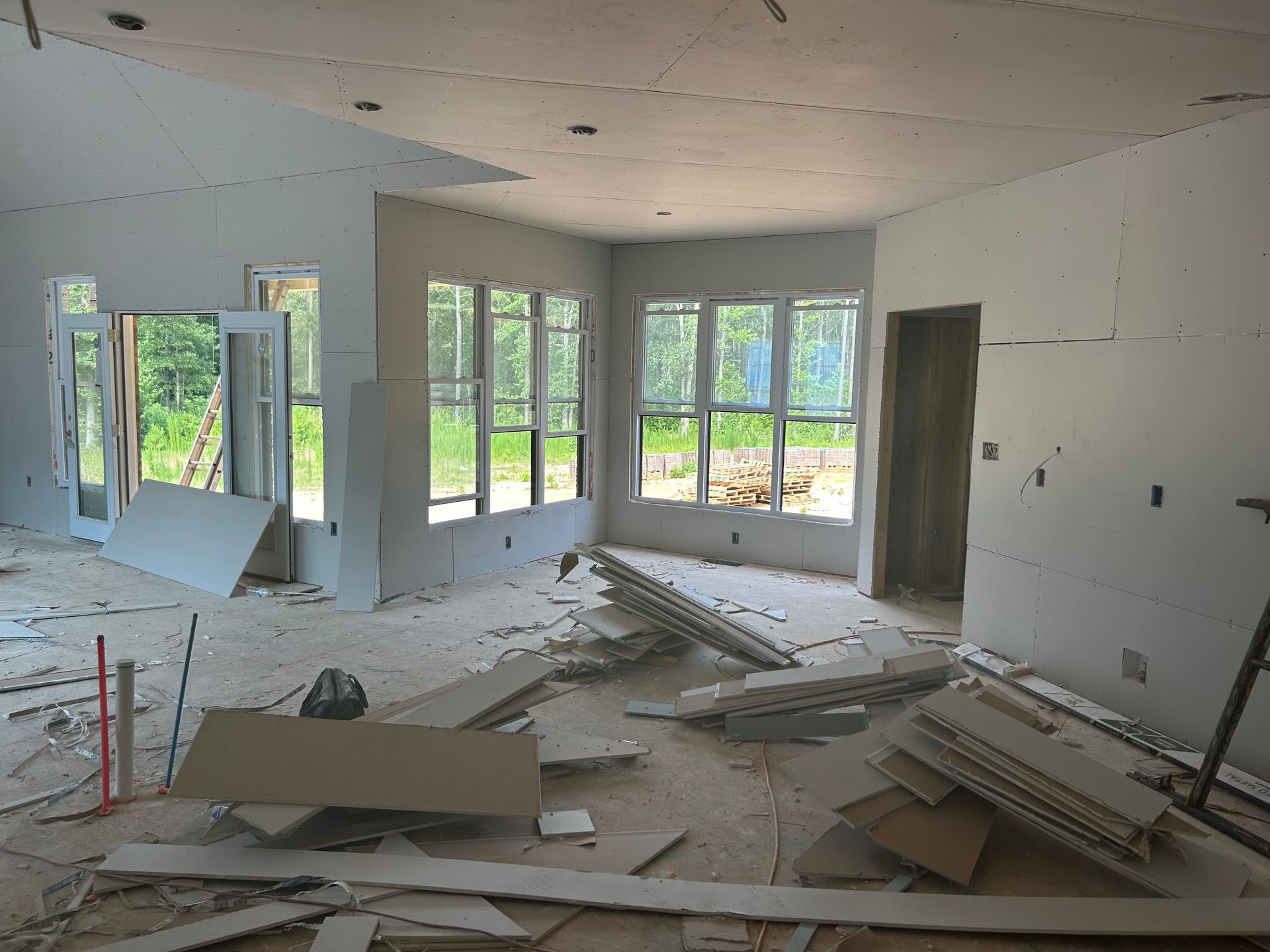 Unfinished room with drywall debris, exposed framing, and large windows letting in daylight.