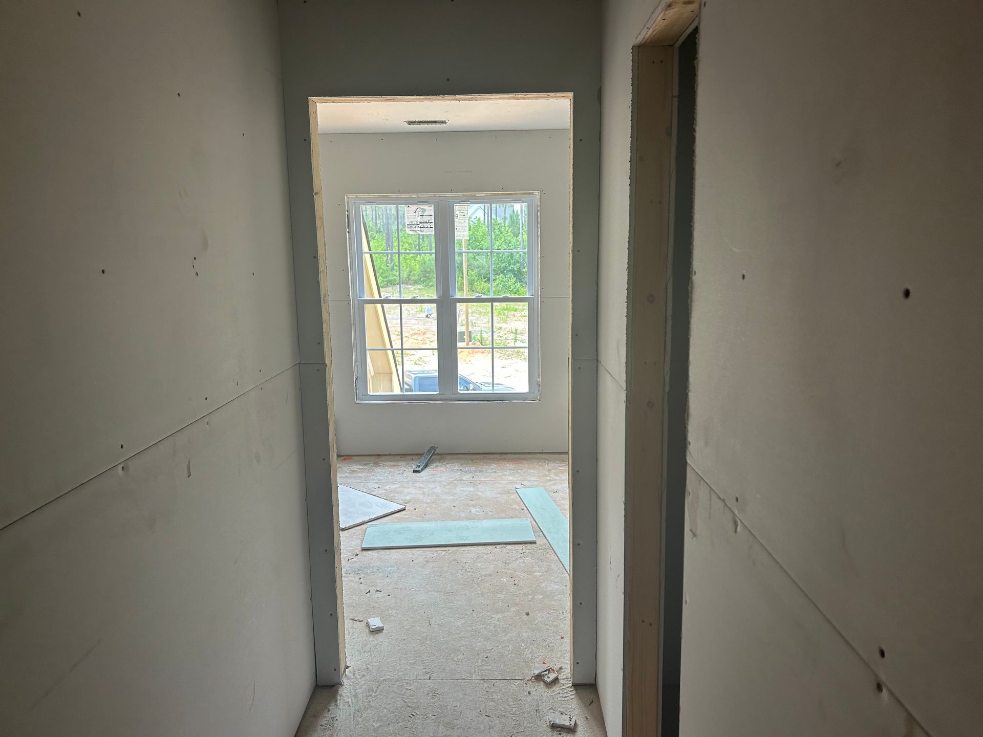 Narrow unfinished hallway with drywall leading to a bright room and window at the end.