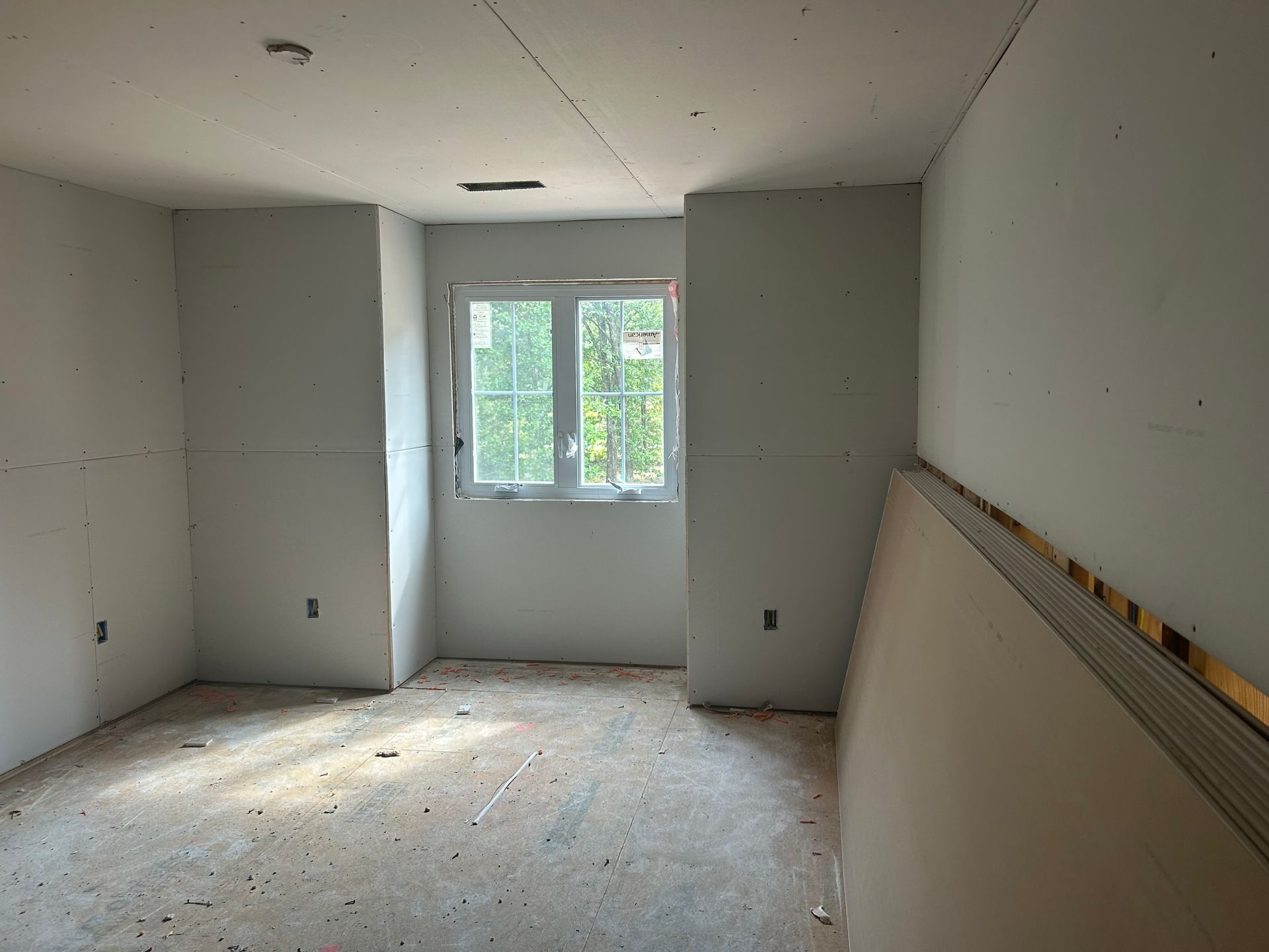 Empty unfinished room with bare drywall, window, and stacked board leaning against the right wall.