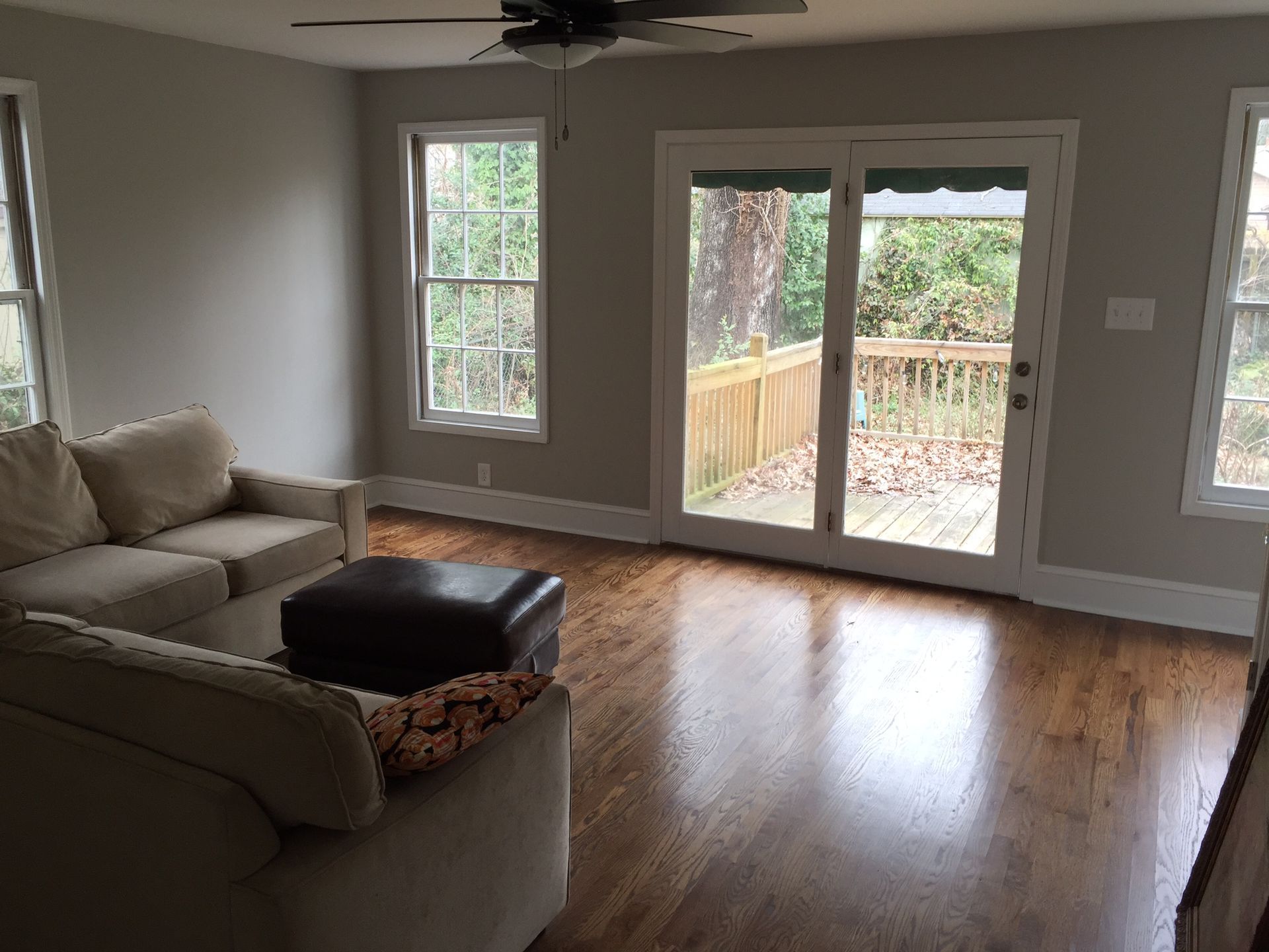 Empty living room with beige sectional, hardwood floors, and sliding glass doors to a fenced backyard.