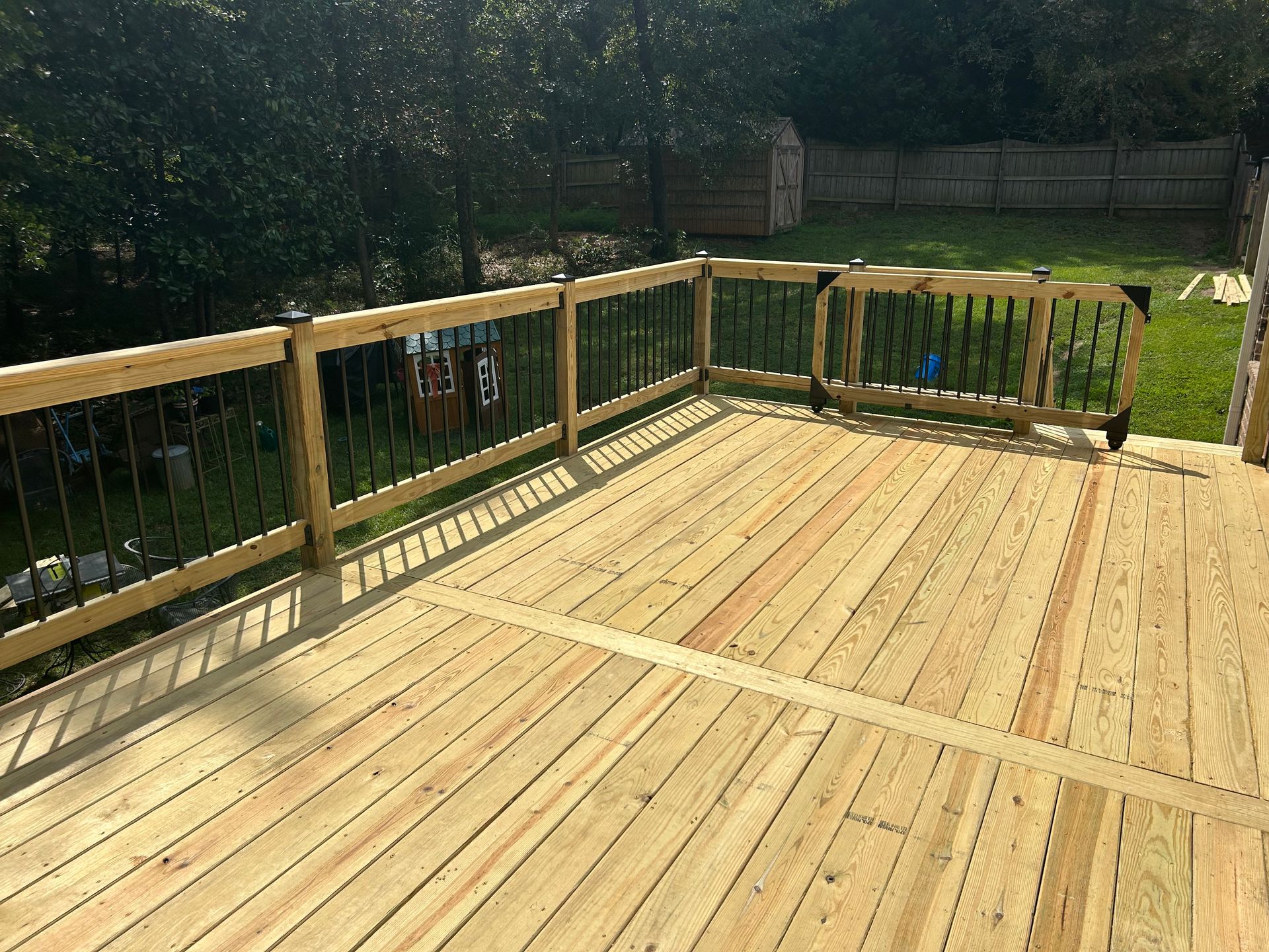 Wooden deck with railing in a sunny backyard, overlooking grass and trees
