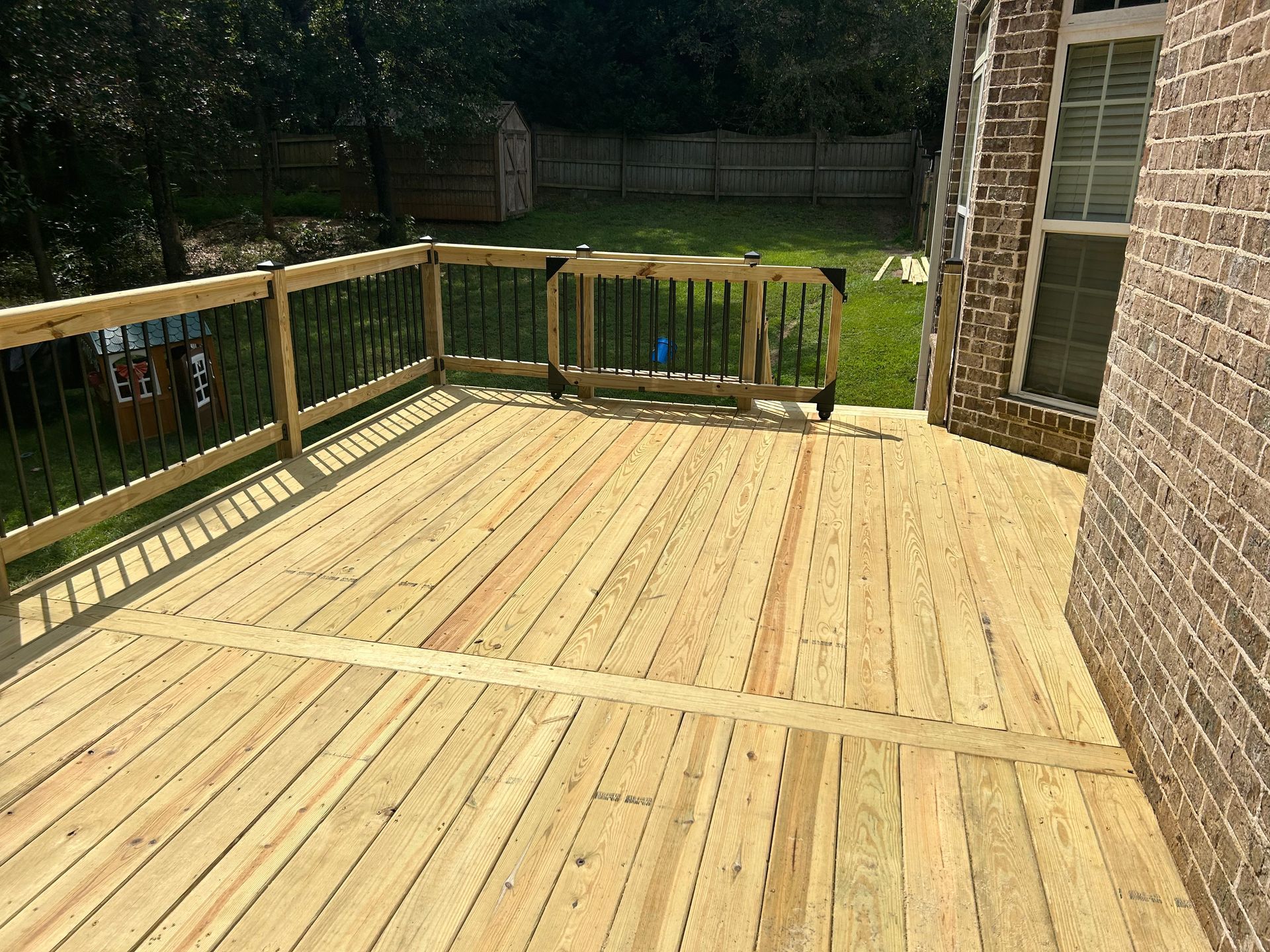 Wooden backyard deck with railing beside a brick house and trees in the background