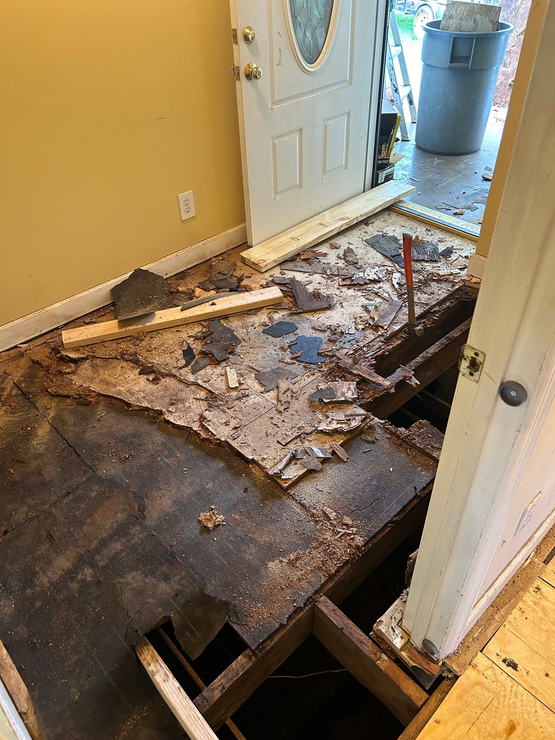 Severe floor damage and exposed joists inside a hallway near an open front door