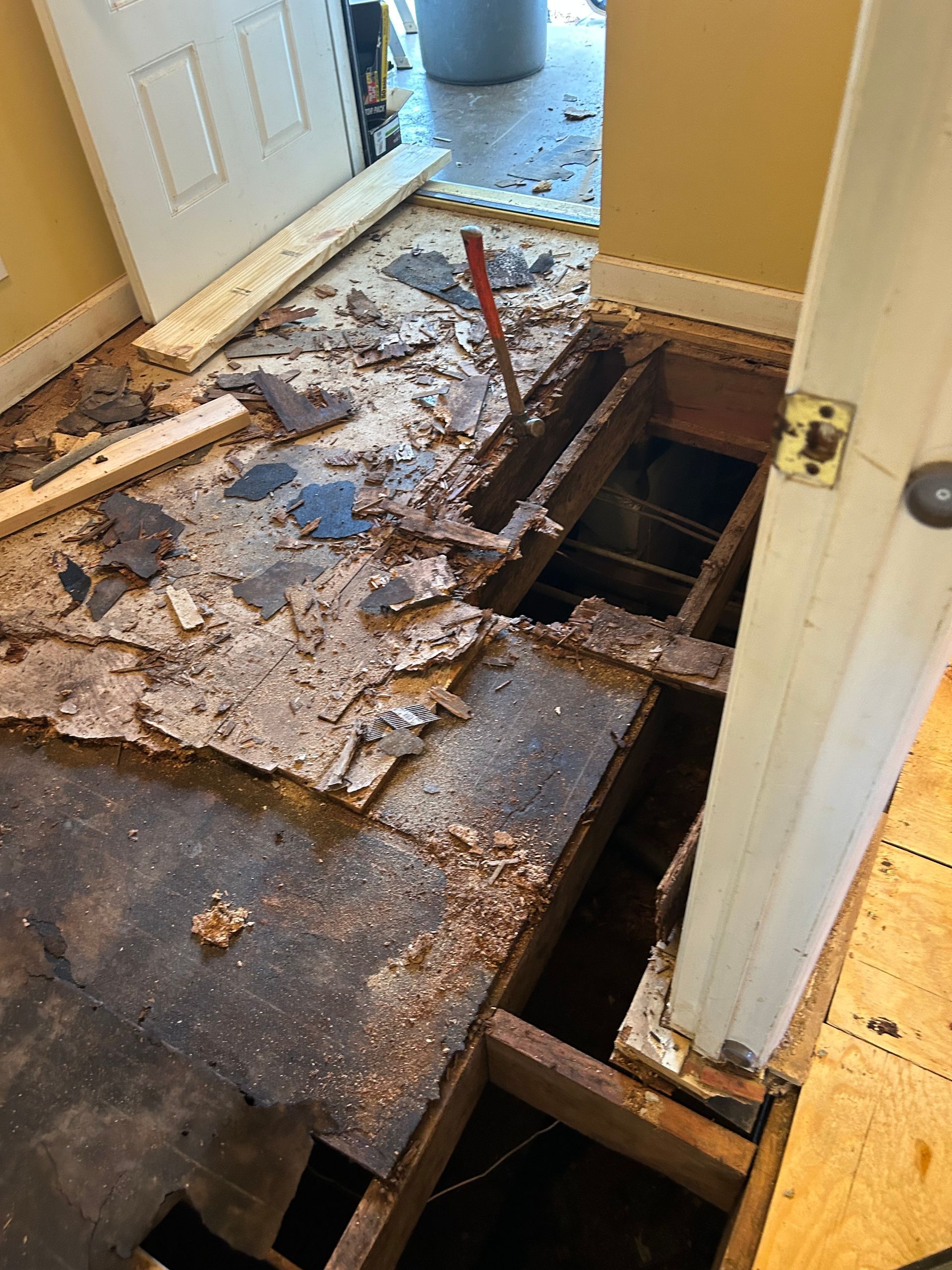 Severely damaged hallway floor with exposed joists, debris, and a large hole near a doorway.