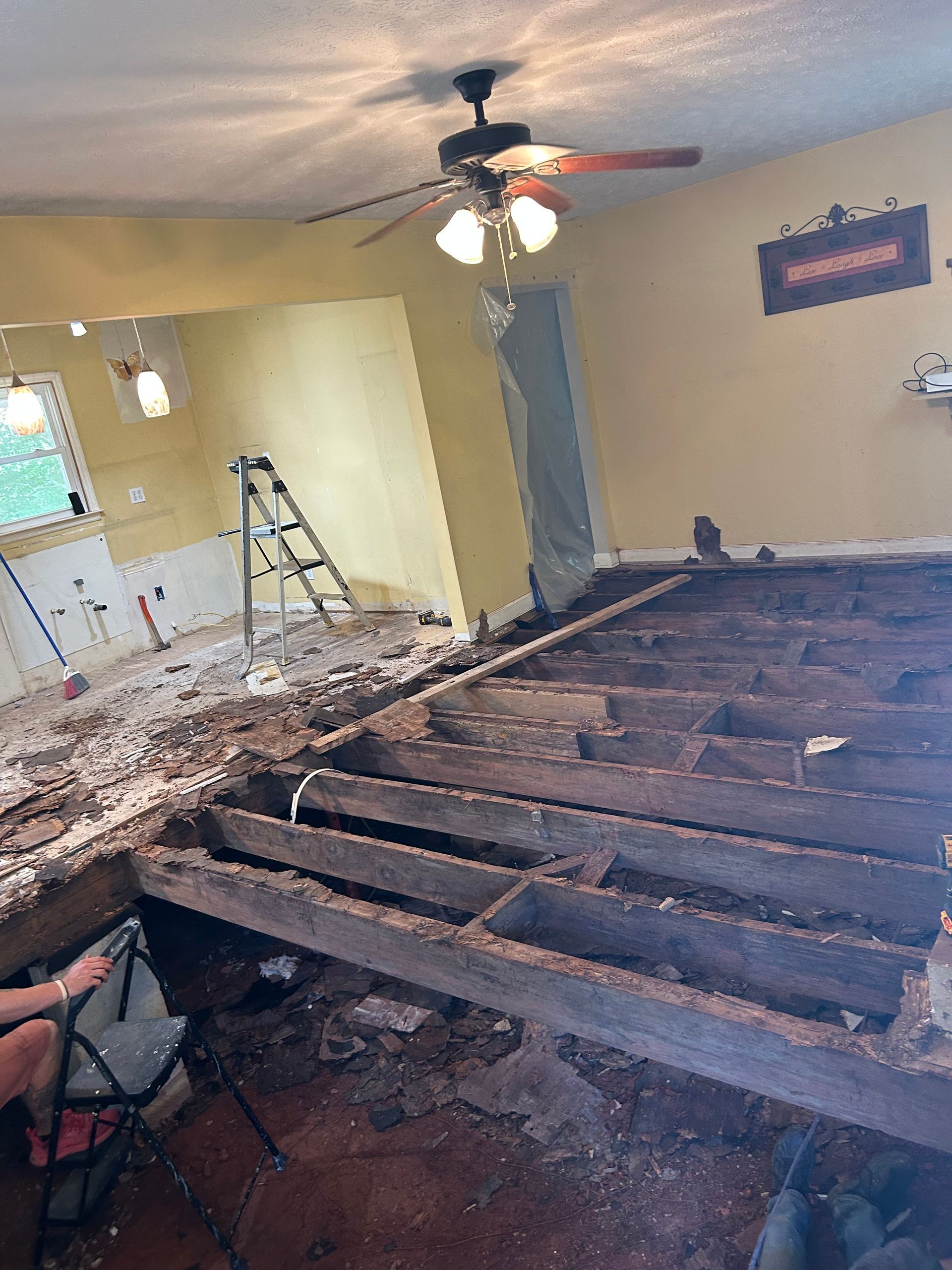 Room under renovation with exposed floor joists, debris, a ladder, and a ceiling fan overhead