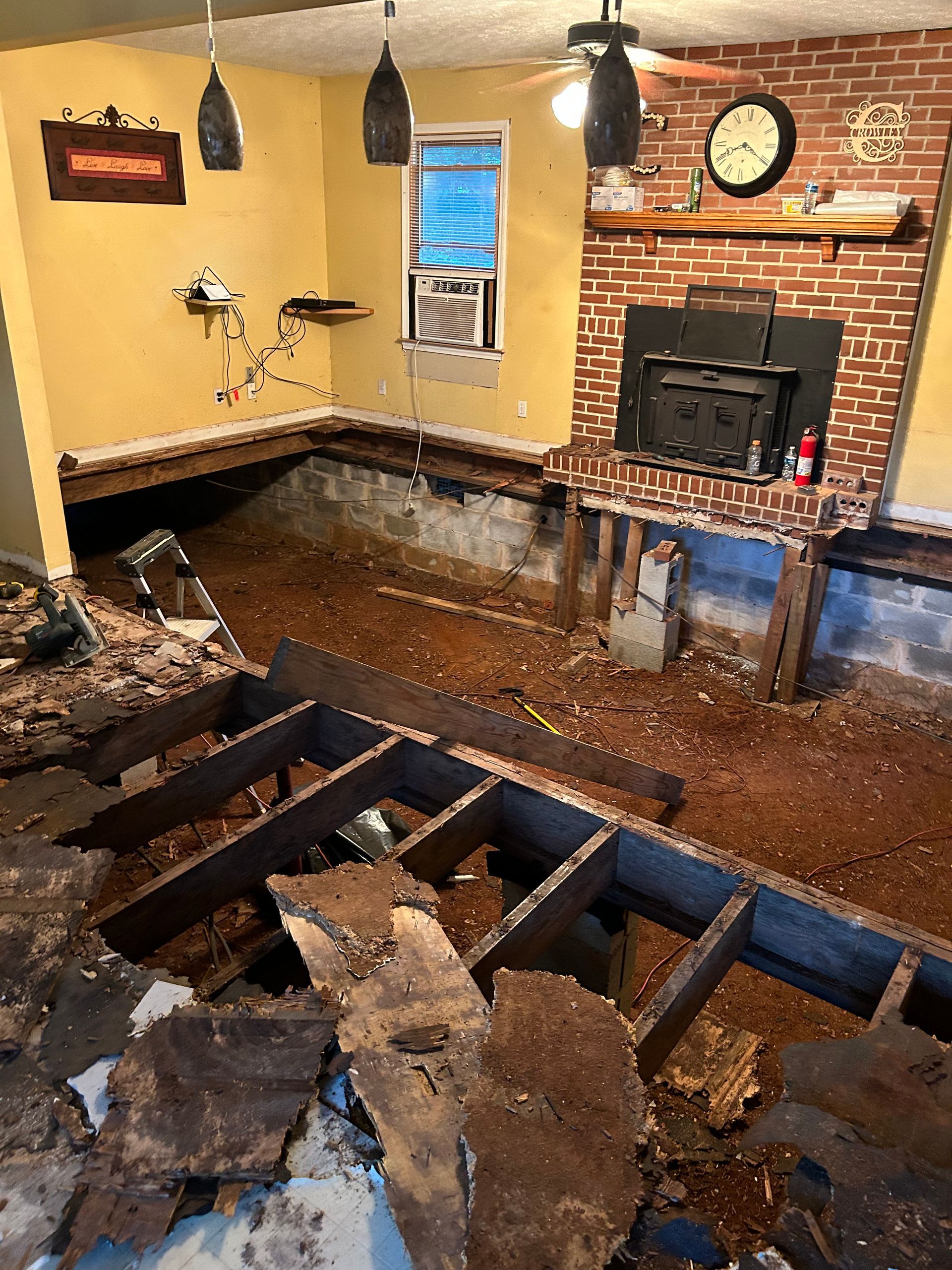 Flood-damaged living room with torn-up floor, exposed joists, wet debris, and a brick fireplace