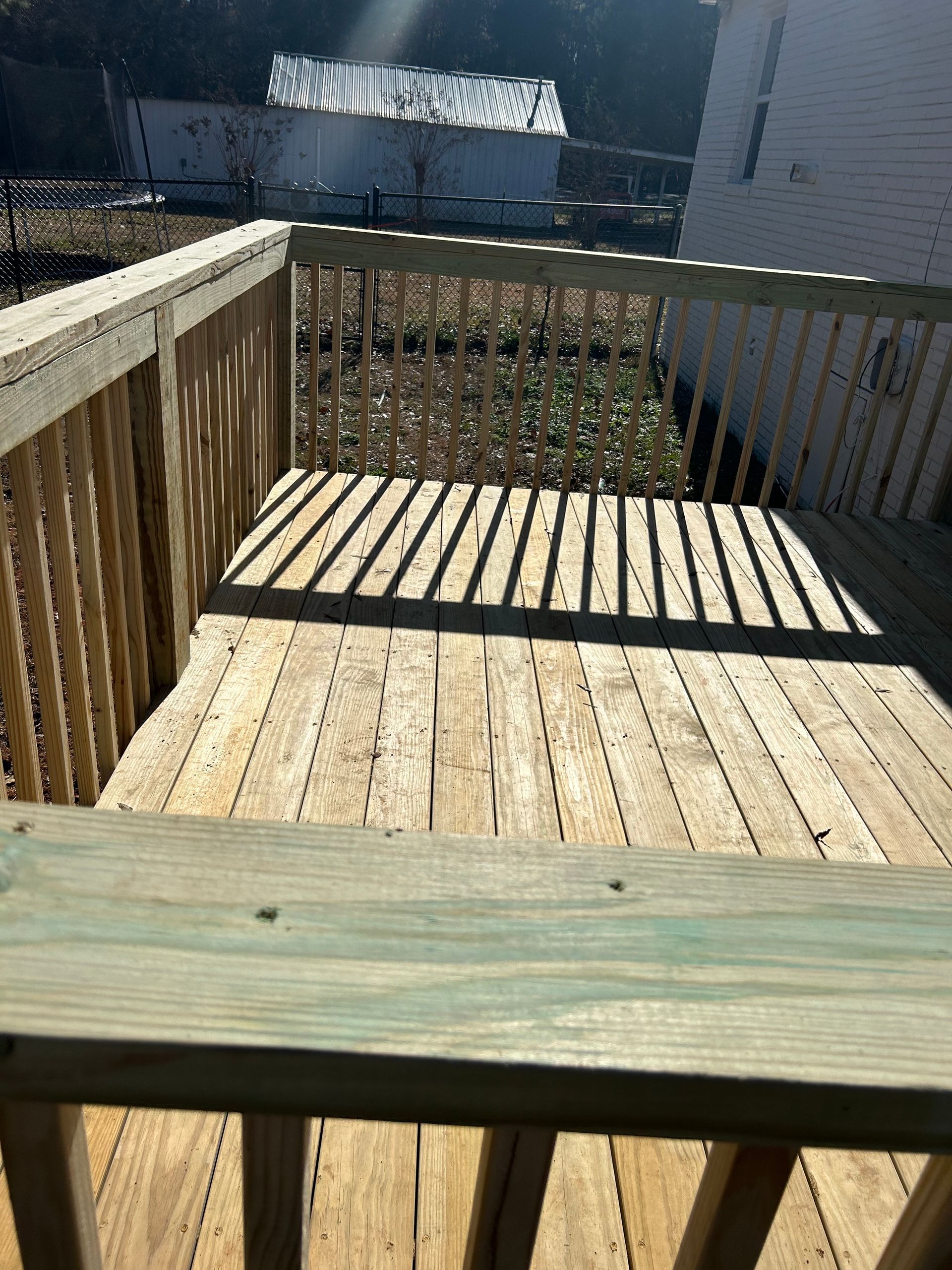 Wooden deck with railings casting shadows in bright sunlight, overlooking neighboring buildings and a fence.