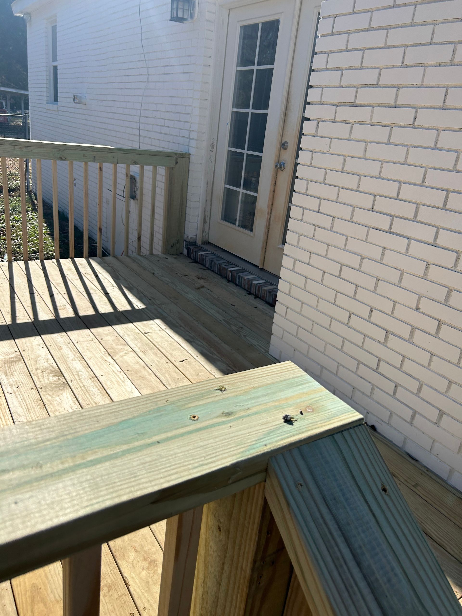 Wooden deck with railing beside a white brick house and glass door, sunlit shadows across the boards