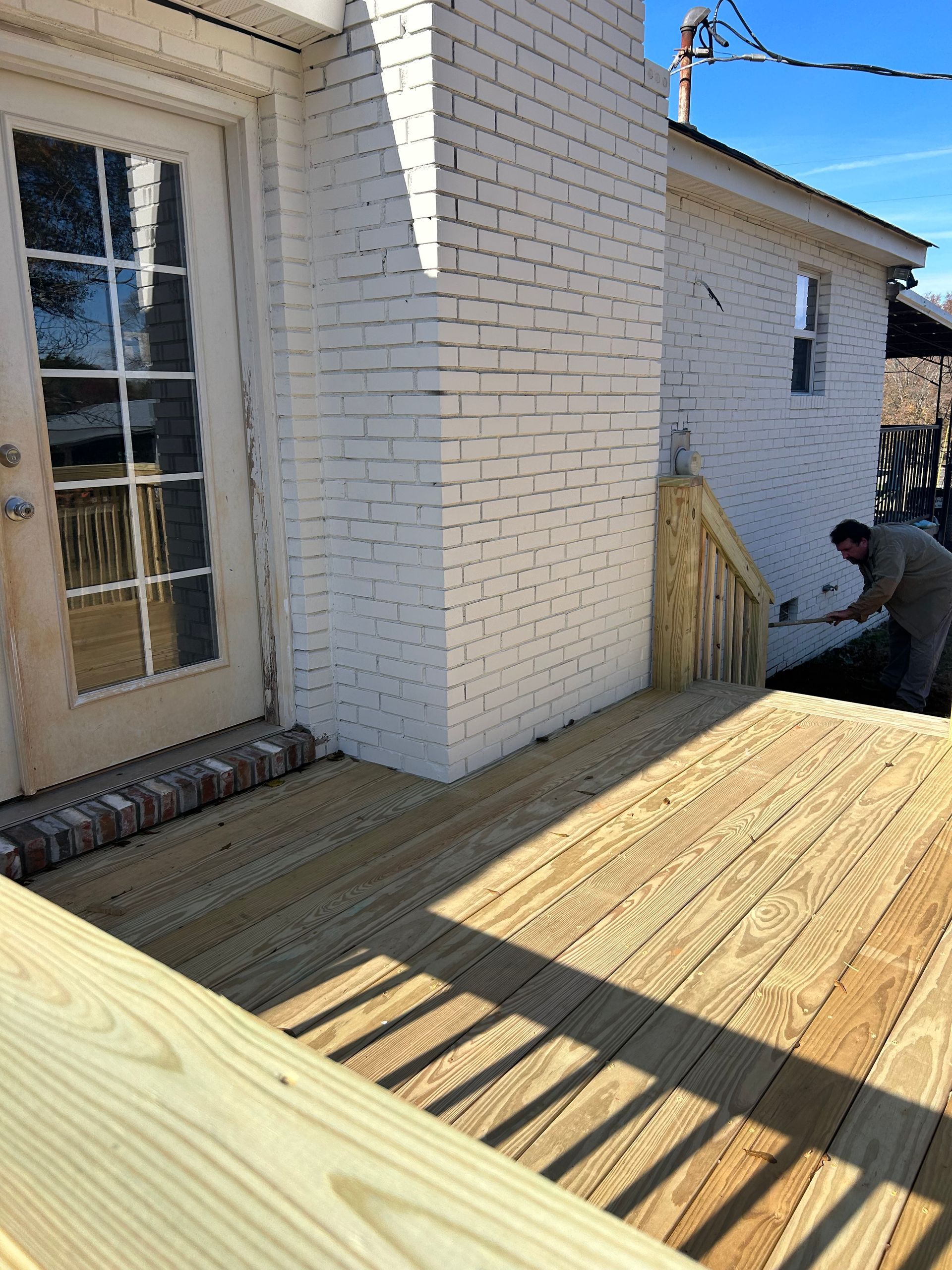 Wooden deck being built beside a white brick house, with a worker kneeling near the corner.