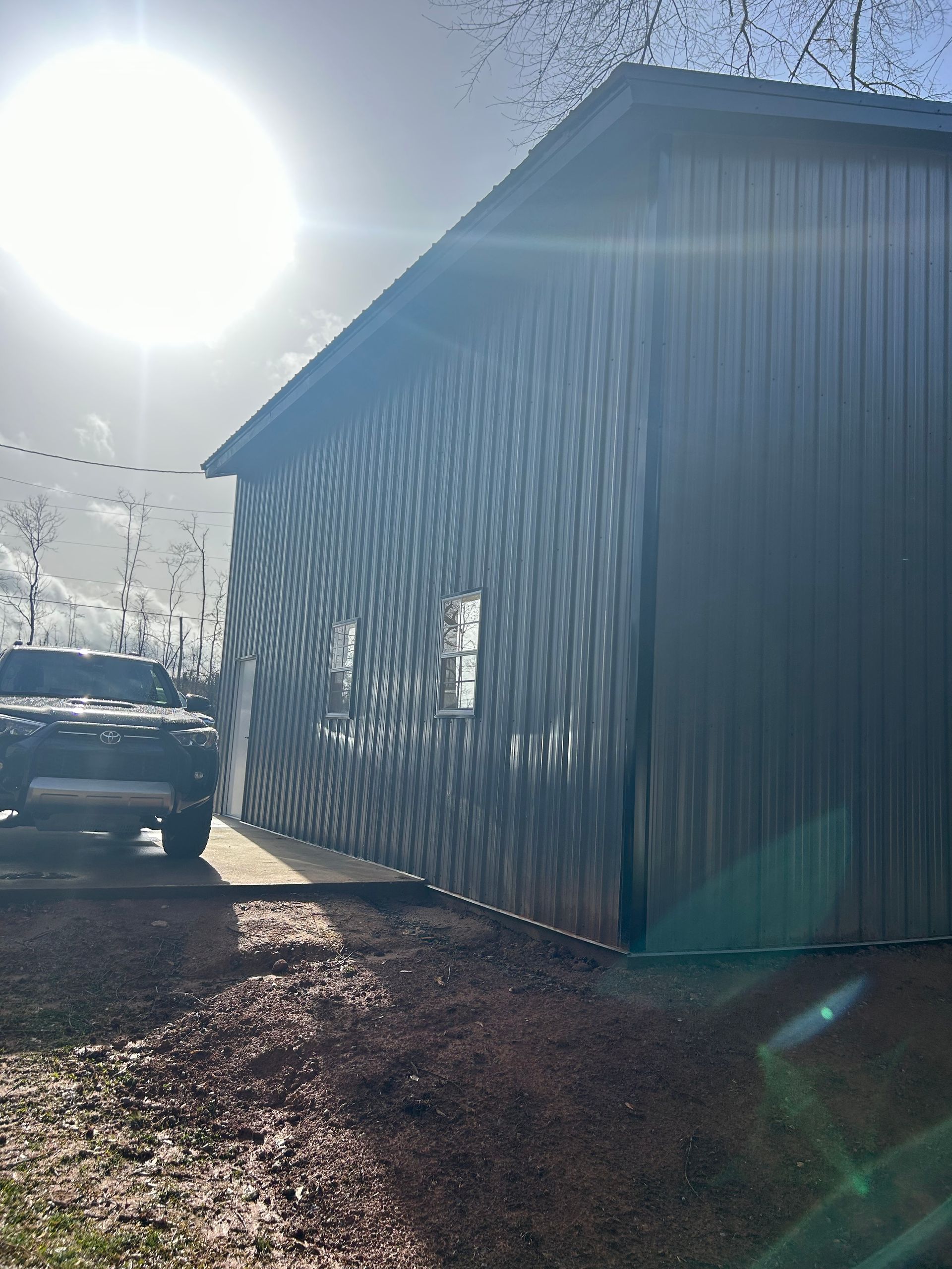Sunny side view of a metal-sided shed with a parked truck beside it on a dirt driveway