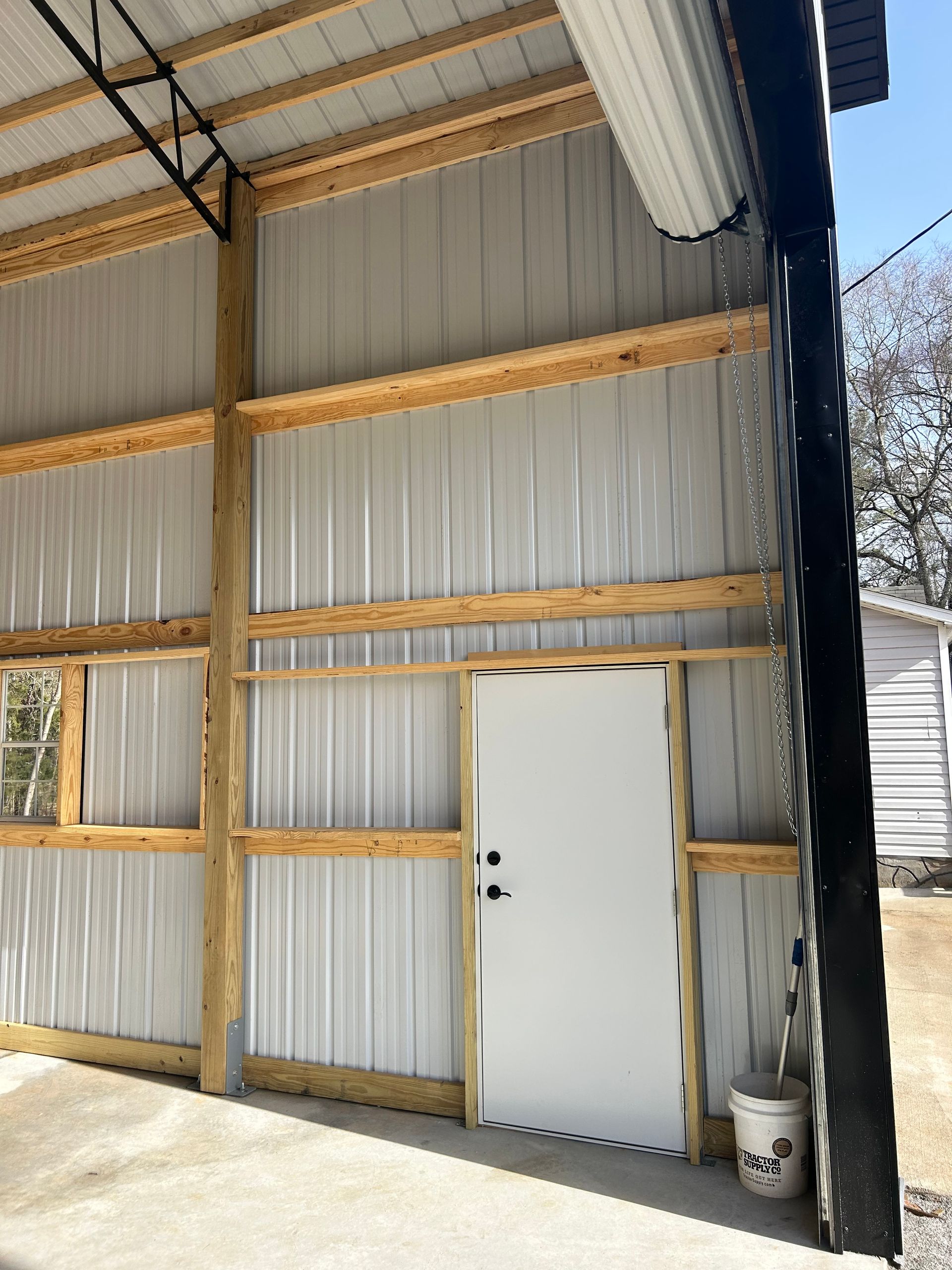 Interior of a metal shed with a white door, corrugated walls, exposed beams, and sunlight on the floor