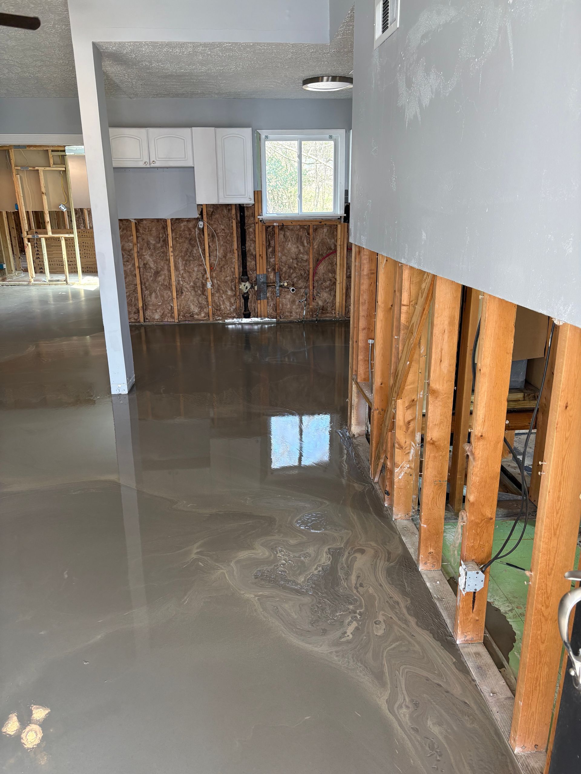 Flooded room with standing water, exposed wall studs, and a kitchen area in the background