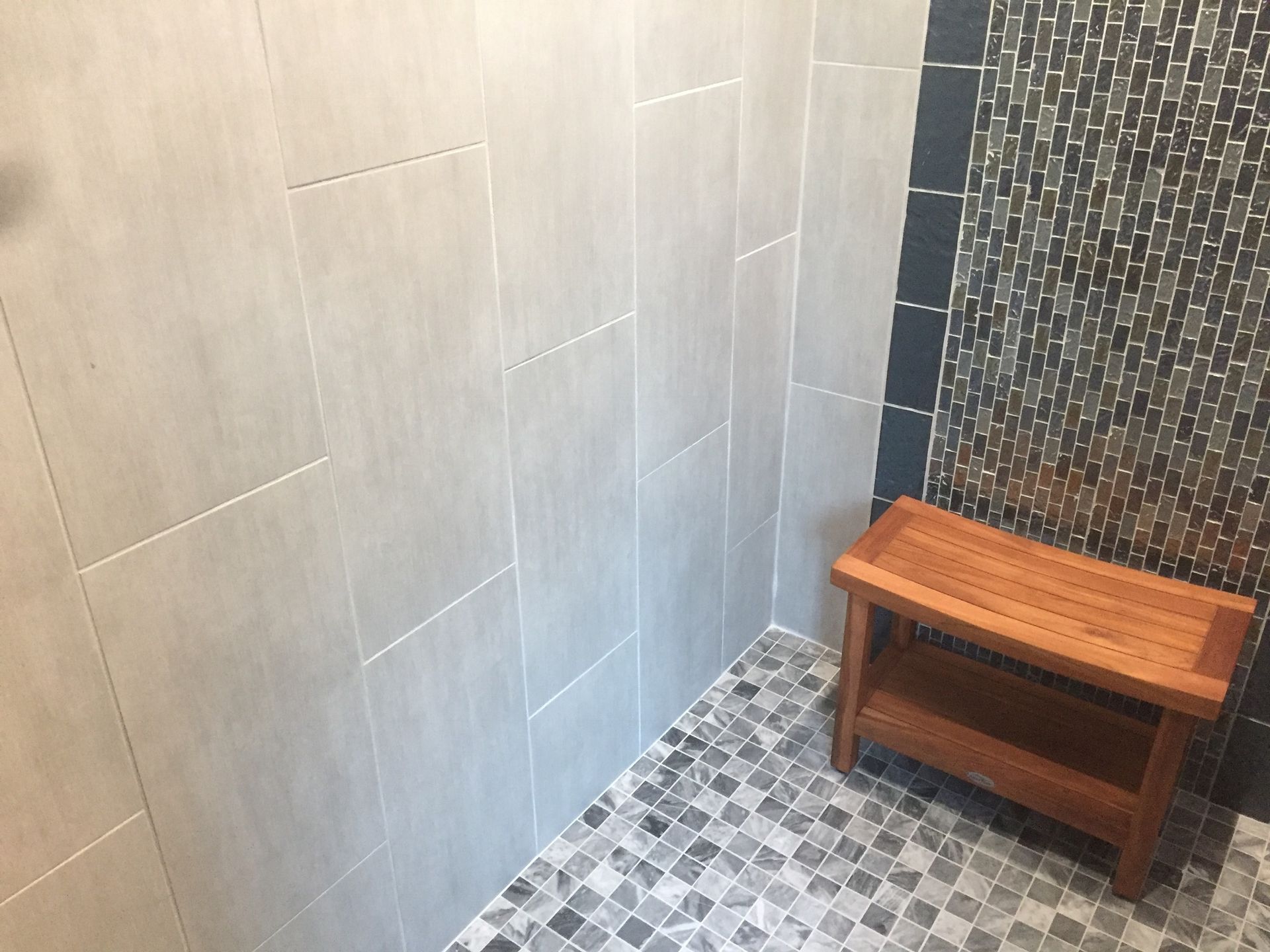 Small tiled shower corner with gray walls, mosaic floor, and a wooden bench stool.