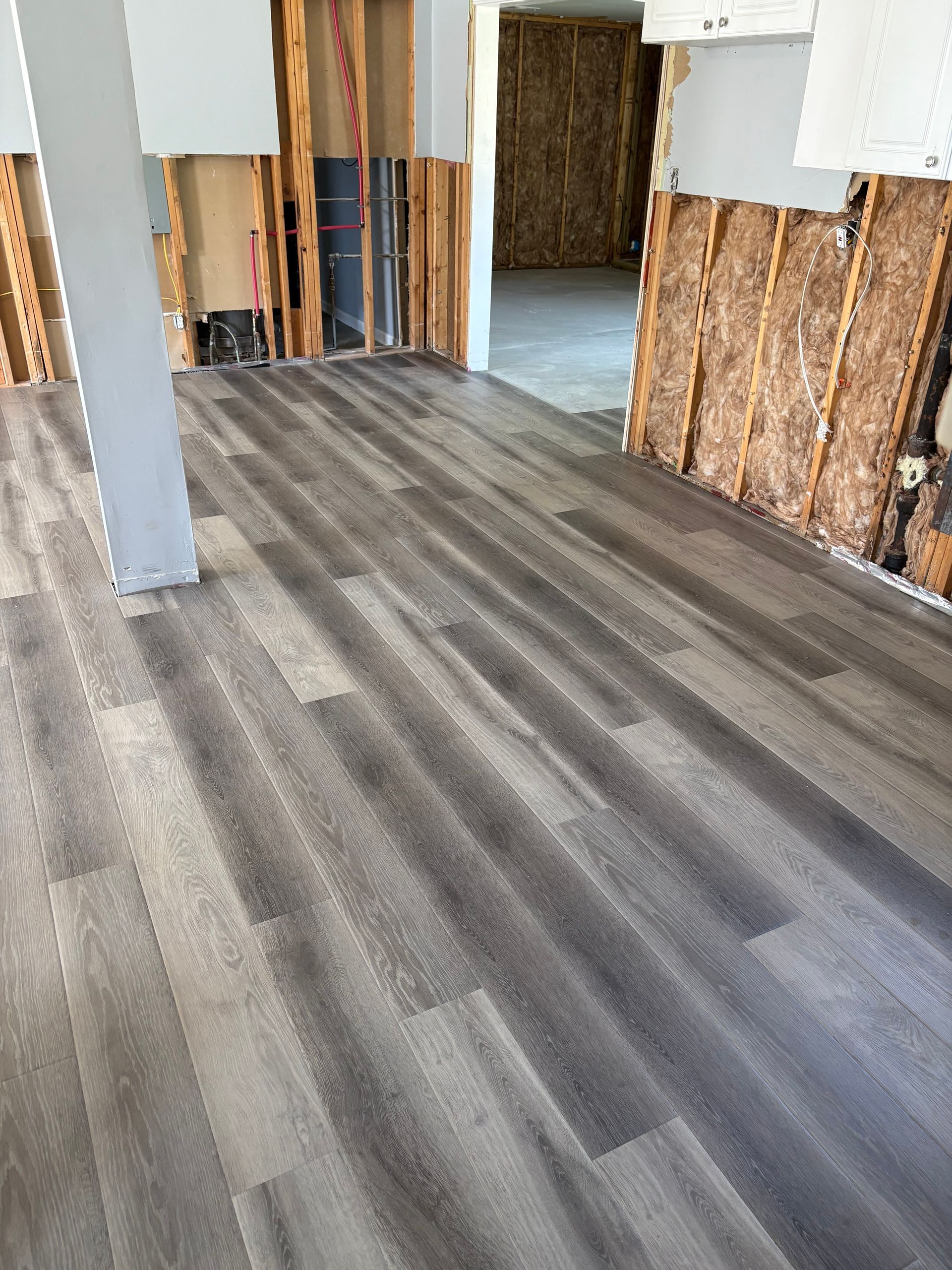 Empty room with gray wood-look flooring, exposed wall studs, and a doorway to another room