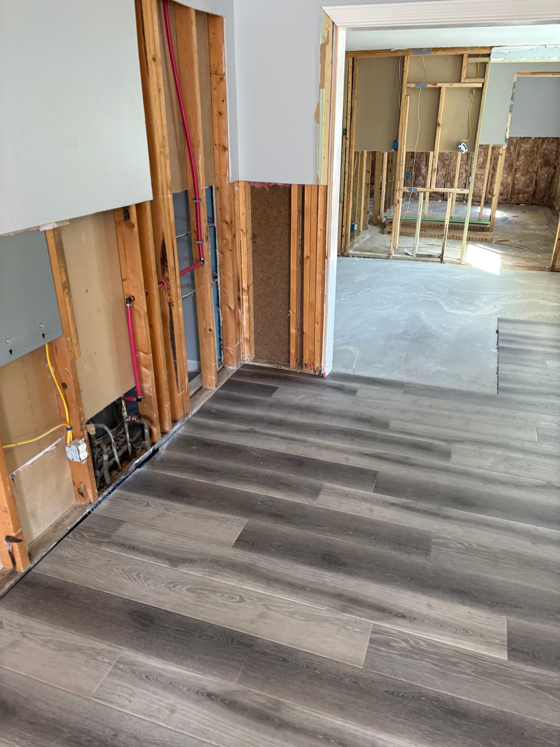 Renovation site with exposed wall studs and gray wood-look flooring leading into an unfinished room